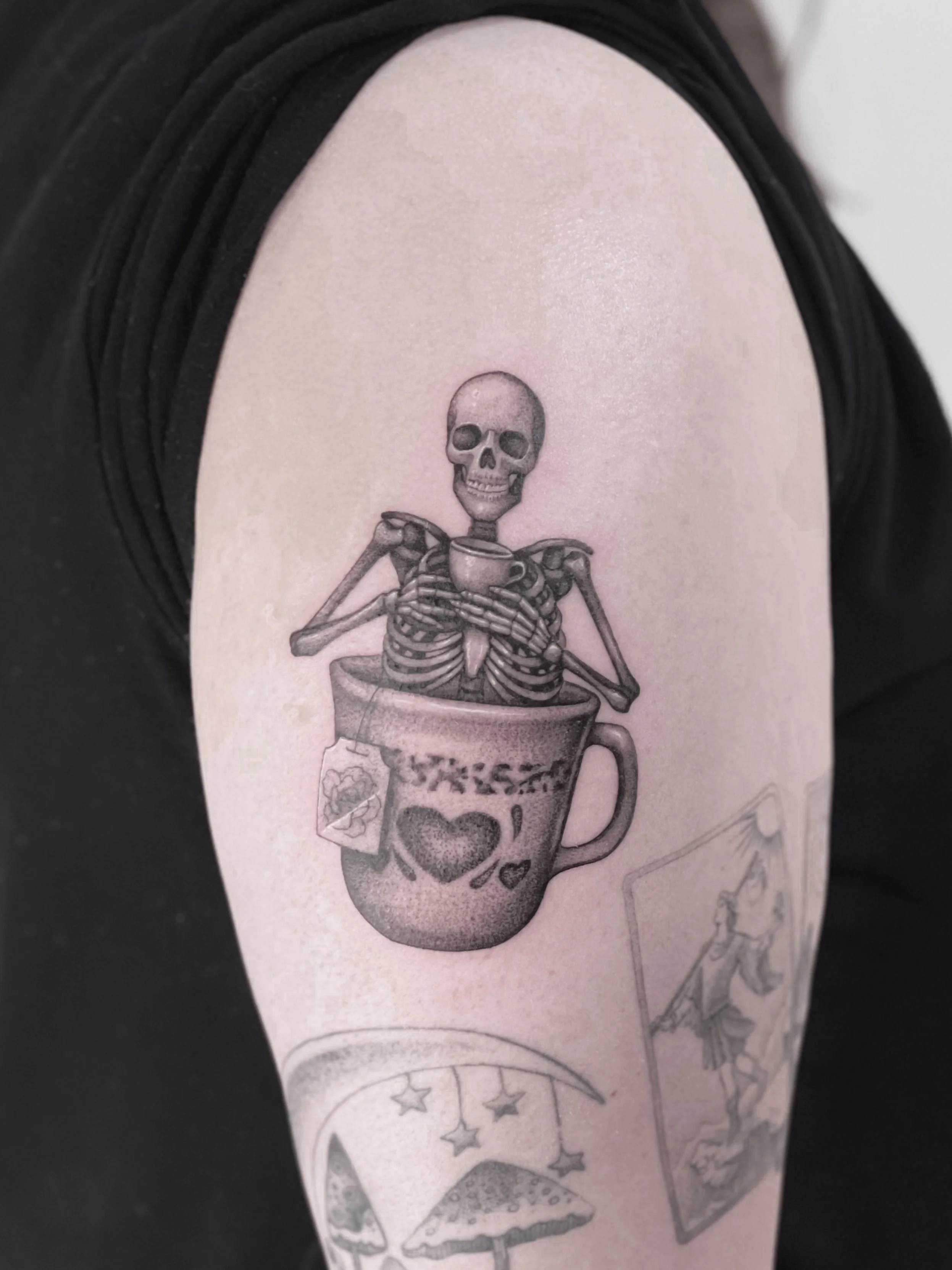 Tattoo of a skeleton holding a cup, sitting inside a large mug with heart and love designs, on a person's arm.