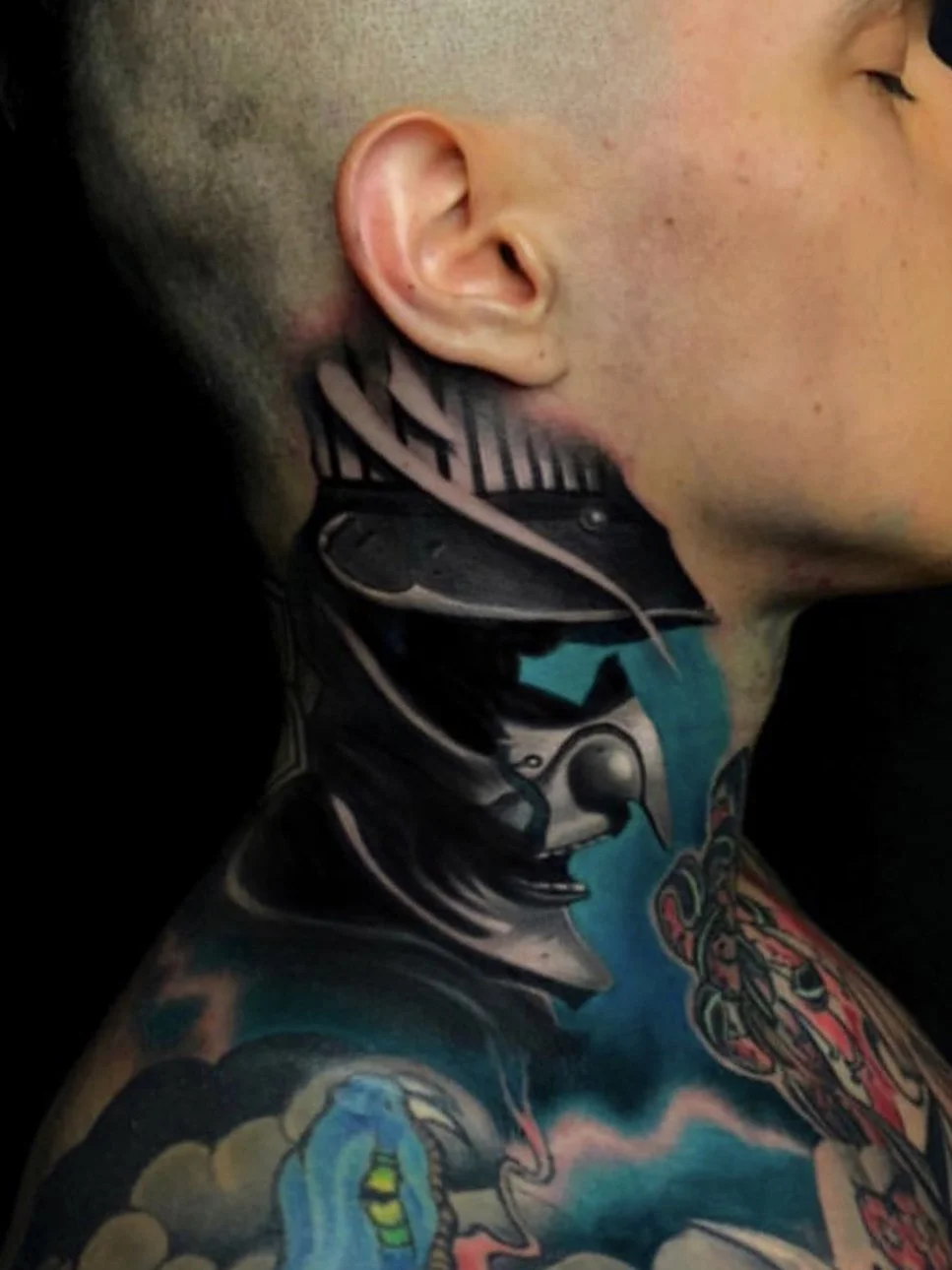 Close-up of a person's face and neck with vibrant, detailed tattoos, including a robotic or armored design on the neck and colorful imagery on the shoulder.