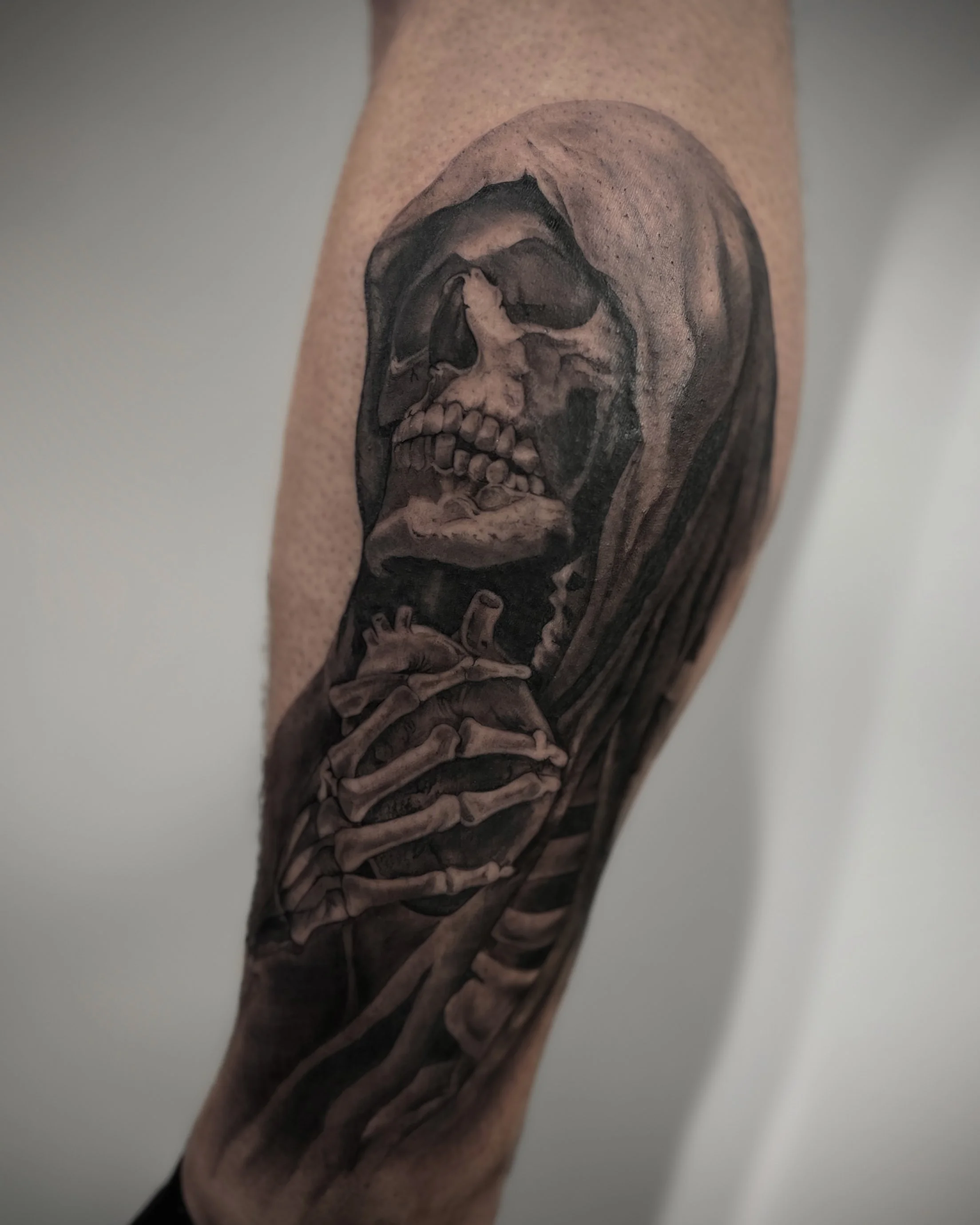 Black and gray tattoo of a skeleton with a hood, holding a smaller skeleton in its hand.