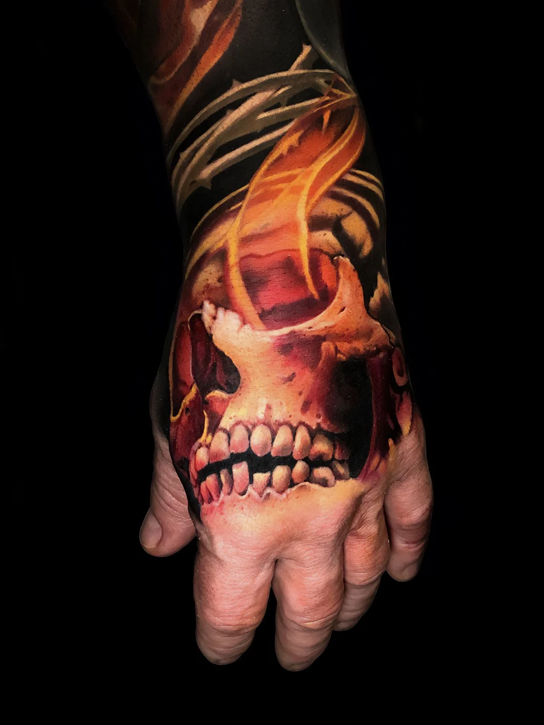 Tattoo of a human skull with flames and smoke details on a person's forearm and hand against a black background.