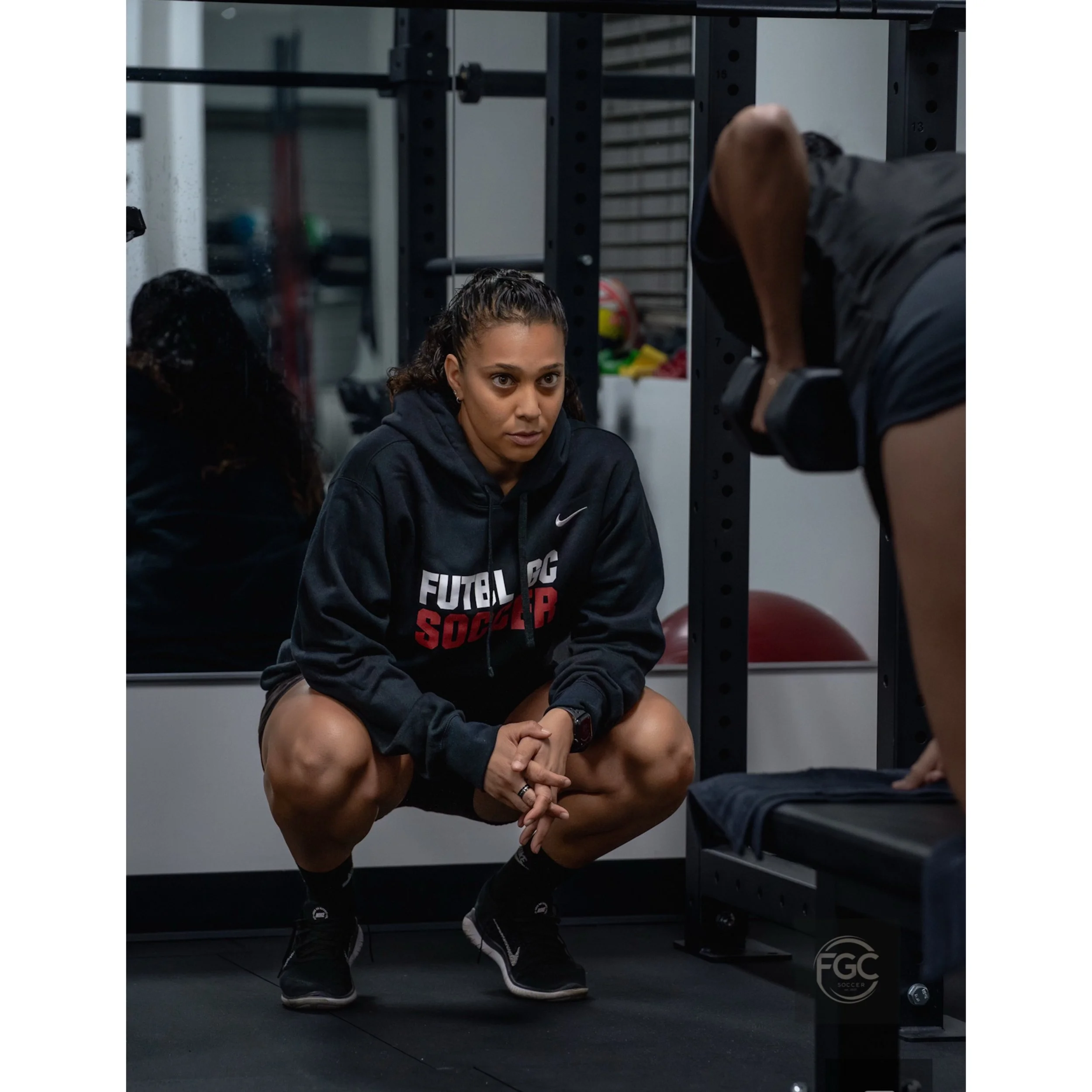 A woman squatting in a gym, wearing a black hoodie with "FUTBOL PC SOCCER" text, observing a person lifting weights.