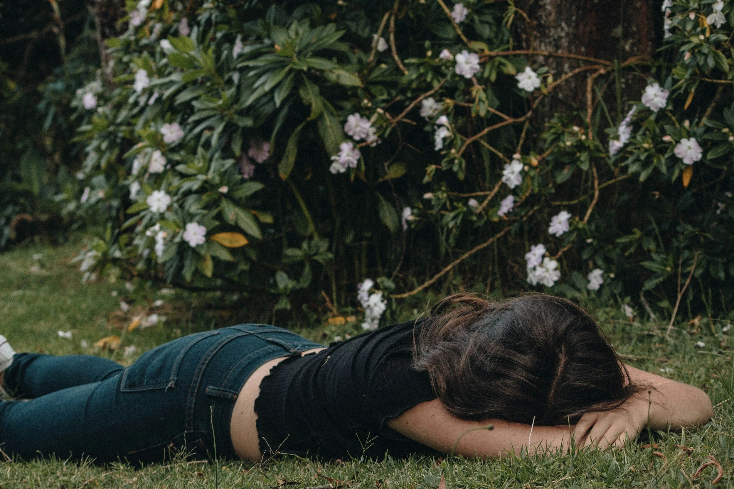 young, petite, white-passing woman with brown hair, wearing black t-shirt and dark navy jeans, lying on the grass face-down on her belly in a garden, with a bush of white flowers in the background