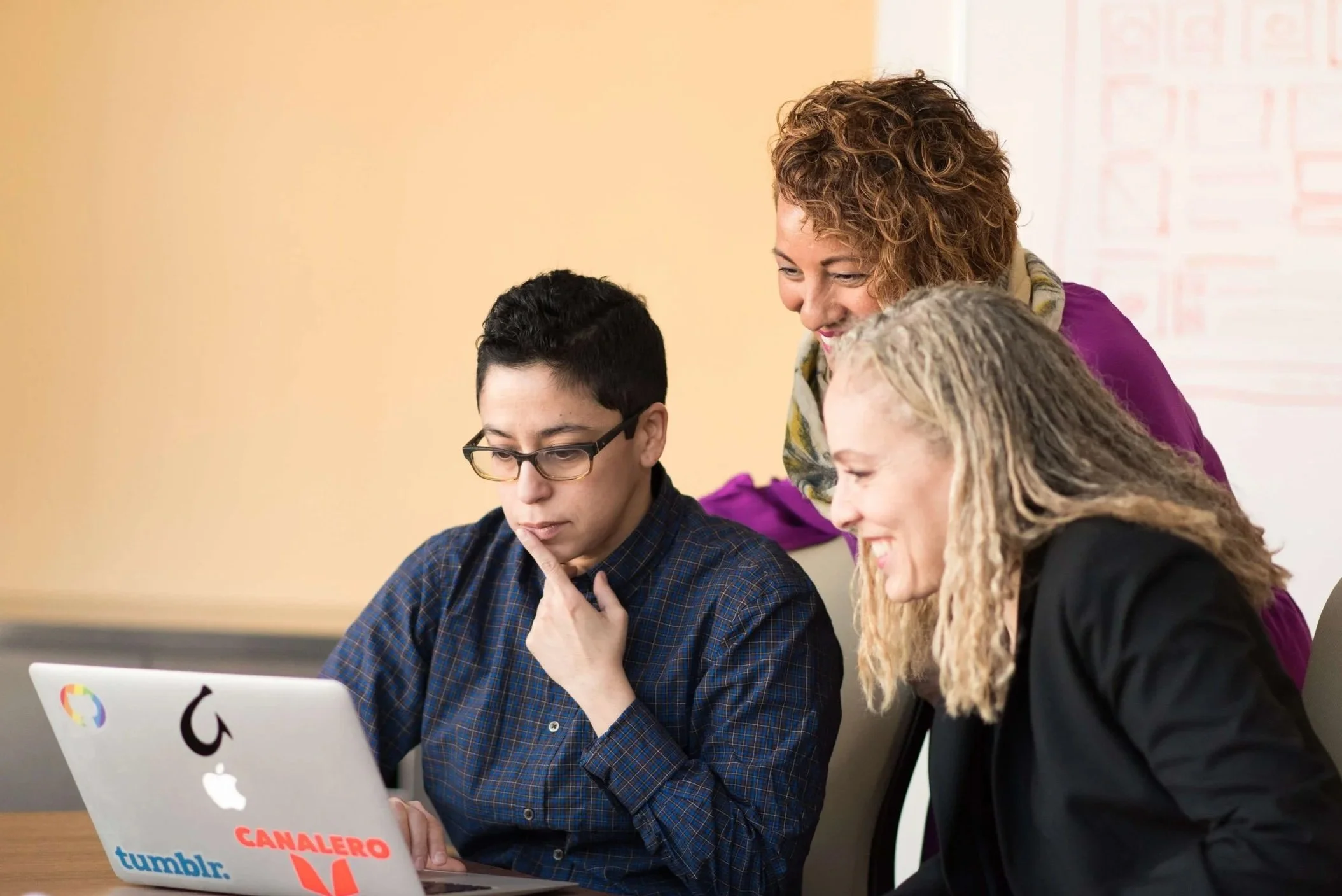 Three racialized gender diverse individuals huddled together and looking at a laptop screen in thought and discussion