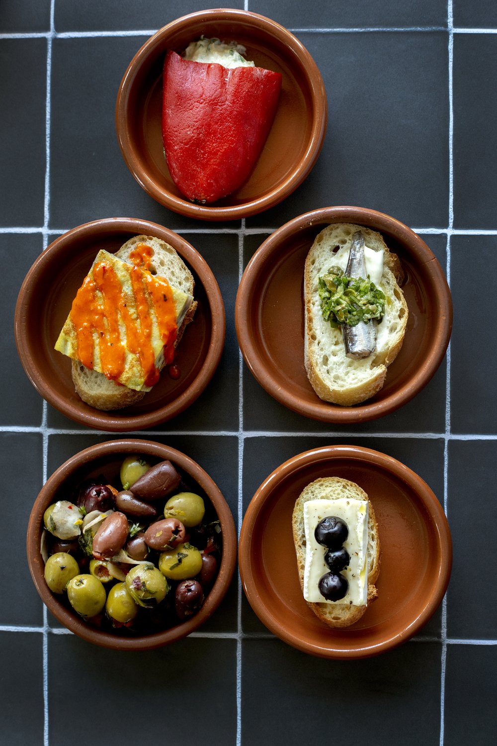 (Top to bottom) Stuffed piquillo pepper, Spanish tortilla with romesco sauce, sardine on toast, marinated olives, and brie with pickled blueberry at Txikiteo