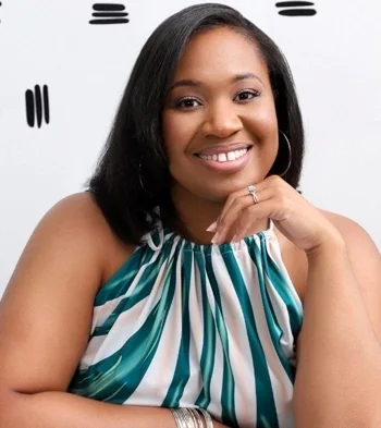 A woman smiling, sitting at a table with her hand resting on her chin.