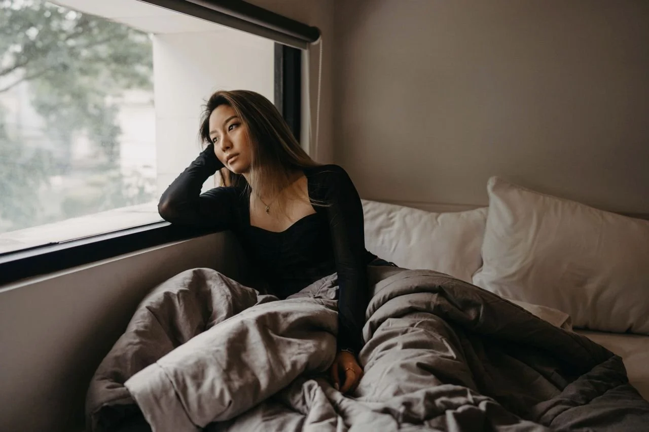 A young woman in a cozy room, pondering by the window, wrapped in blankets