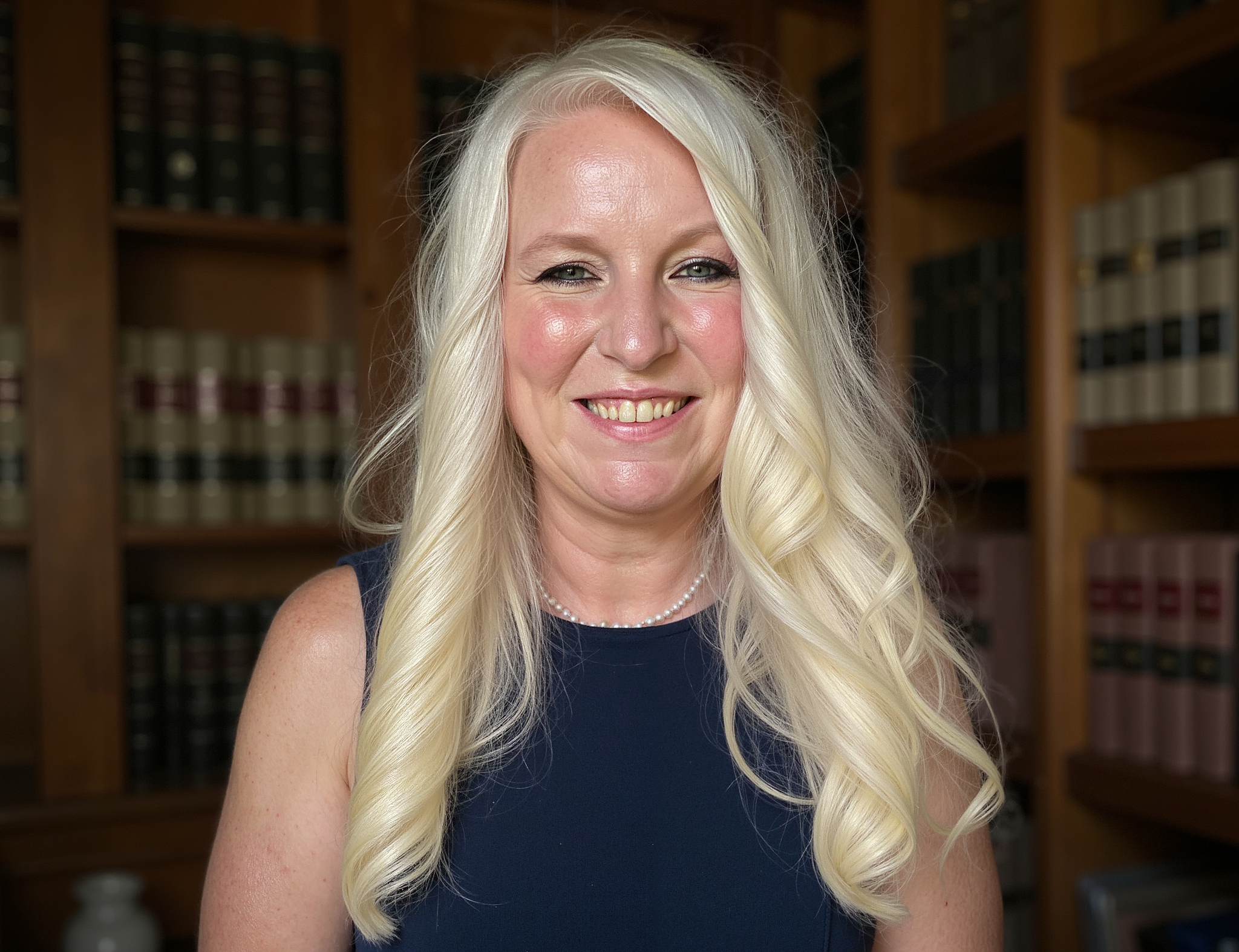 A smiling blonde woman with wavy hair wearing a navy blue sleeveless dress and a pearl necklace, standing in front of a bookshelf filled with law books.