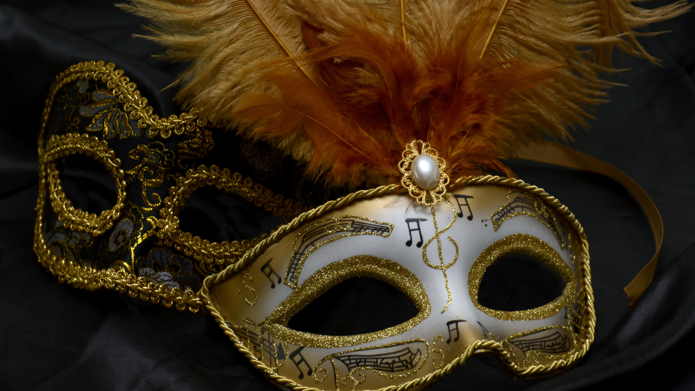 A decorative gold and black masquerade mask with musical notes design, gold trim, and feathers on a black background.