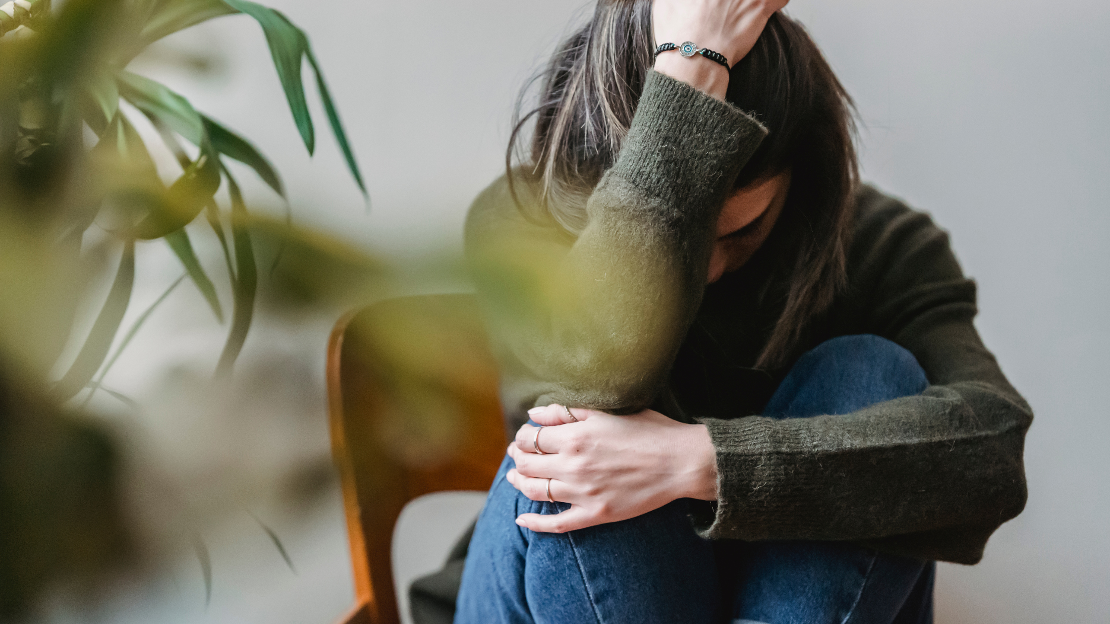 A woman sitting with her head bowed and hand on her forehead, wearing a dark sweater and blue jeans, sitting on a wooden chair, with some green leaves partially obscuring the view in the foreground.