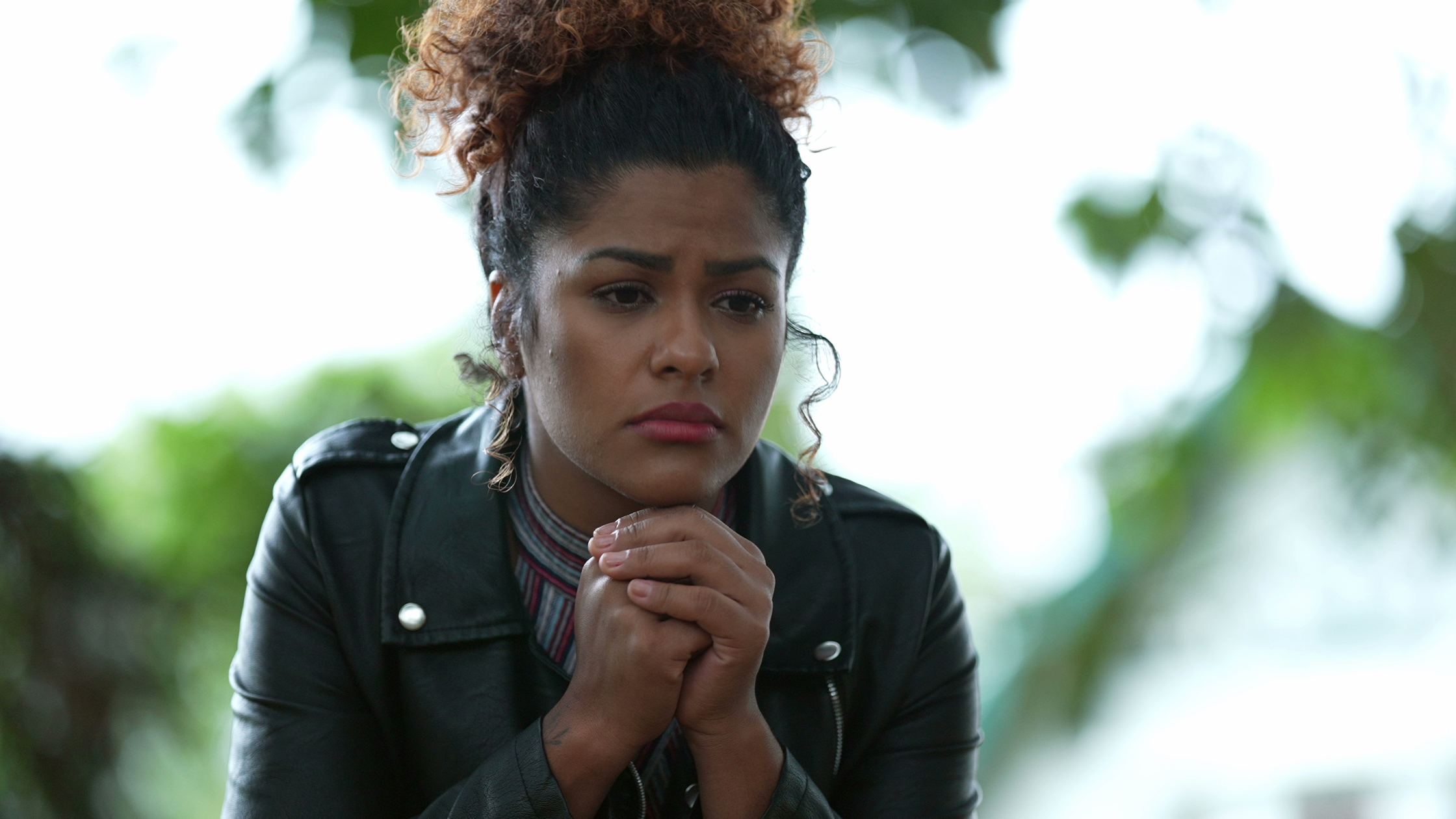 A woman with curly hair tied up in a bun, wearing a black leather jacket, cups her hands together and looks down thoughtfully against a blurred background of green foliage.