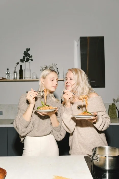 Two women who look related enjoying pasta together in a kitchen.