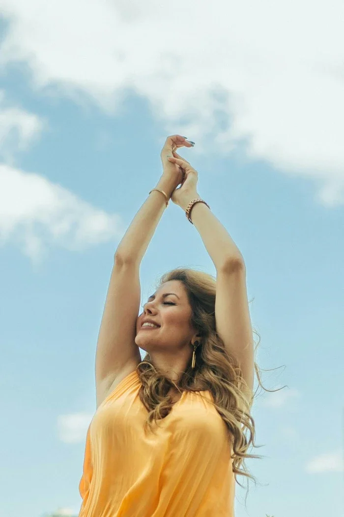 A woman smiling with her hands up in front of a blue sky.