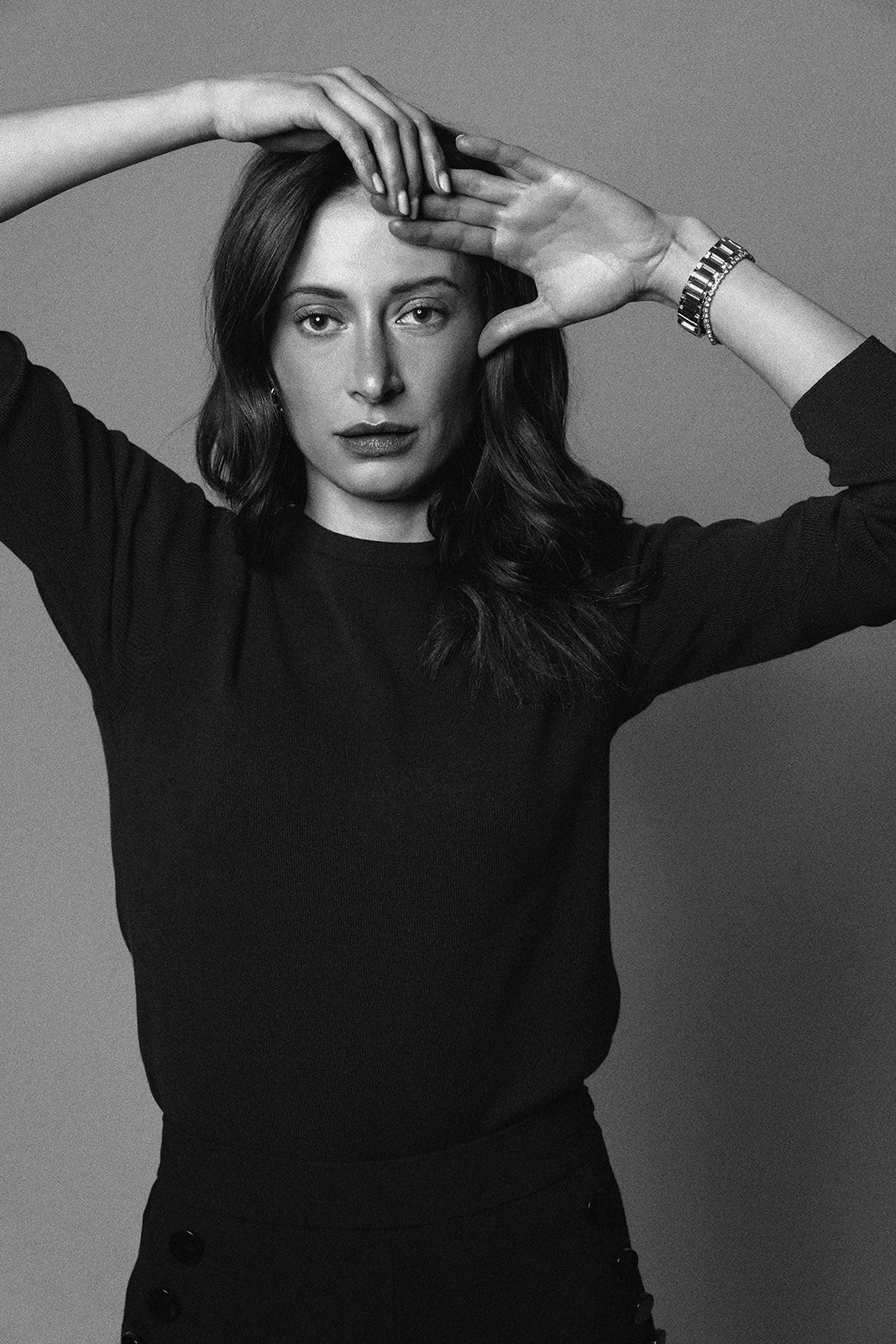 Black and white photo of a woman with dark wavy hair, wearing a black long-sleeve top, looking directly at the camera with one hand on her forehead and the other framing her face, wearing a watch on her wrist.