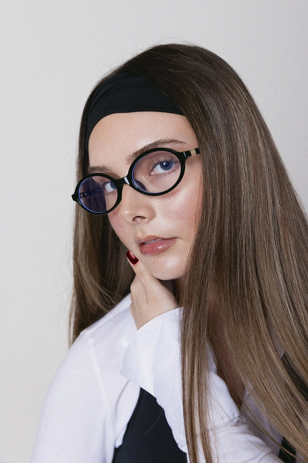 A woman with long brown hair wearing black-rimmed glasses, a black headband, and a white shirt with a black top underneath. She is posing with her hand near her face.