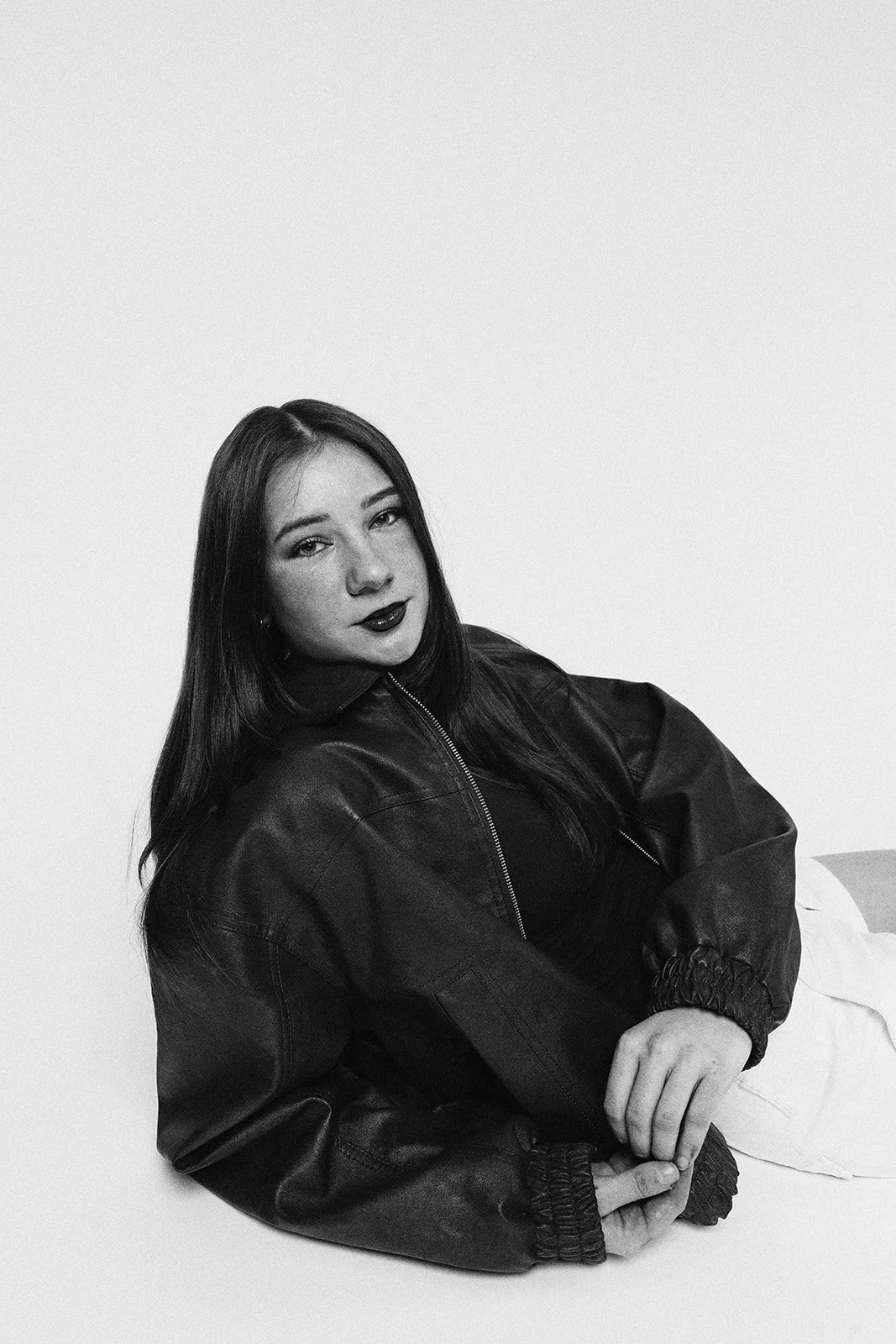 A black and white photo of a woman with long dark hair, wearing a leather jacket, sitting against a plain background.