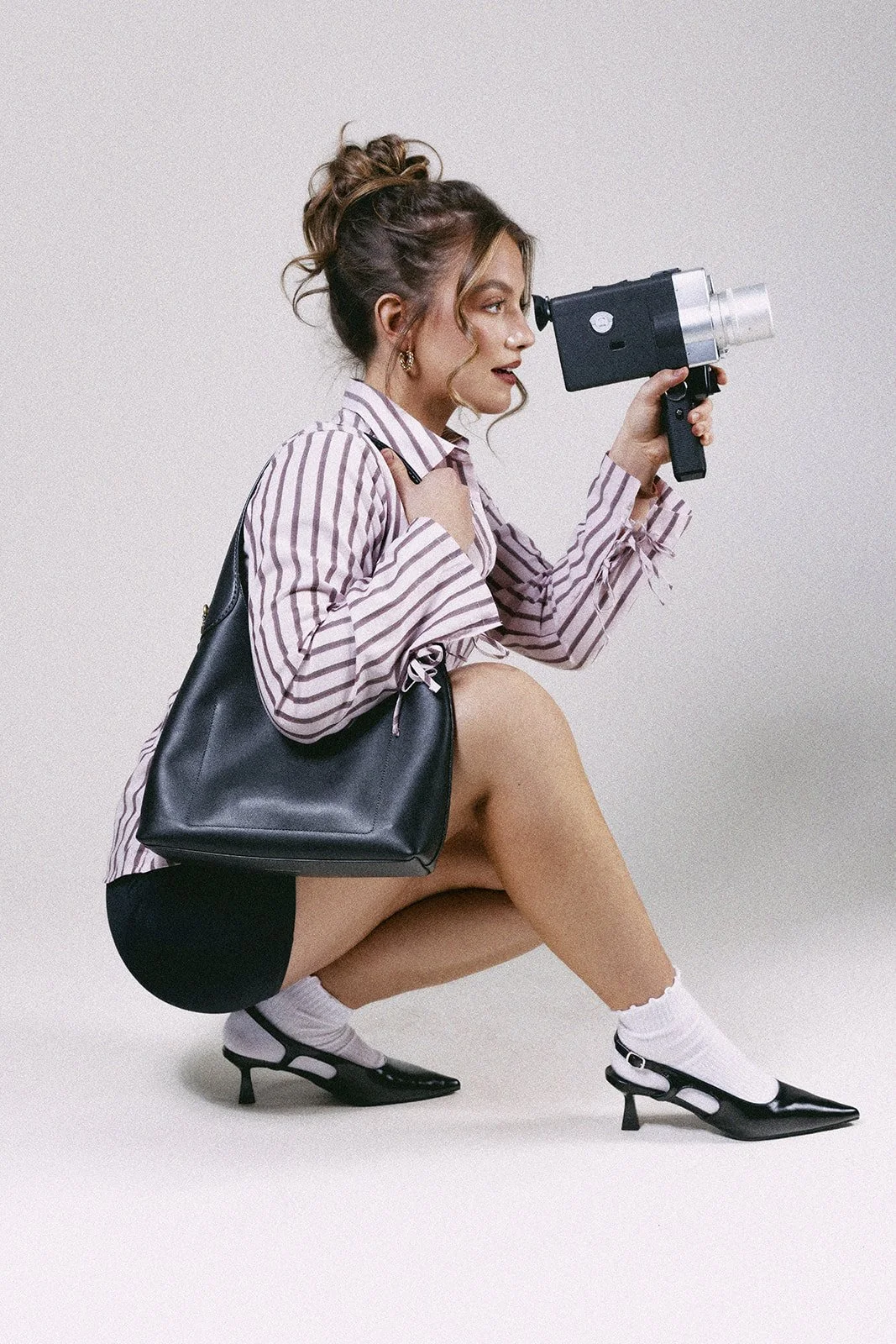A woman squatting with one knee up, holding a vintage camera to her face, wearing a striped shirt, black shorts, white socks, and black high heels, with a black handbag over her shoulder, against a plain light background.