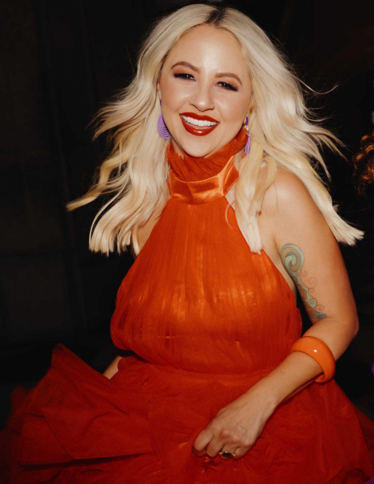 A woman with blonde hair, red lipstick, and purple earrings, wearing an orange dress, smiling and winking at the camera.