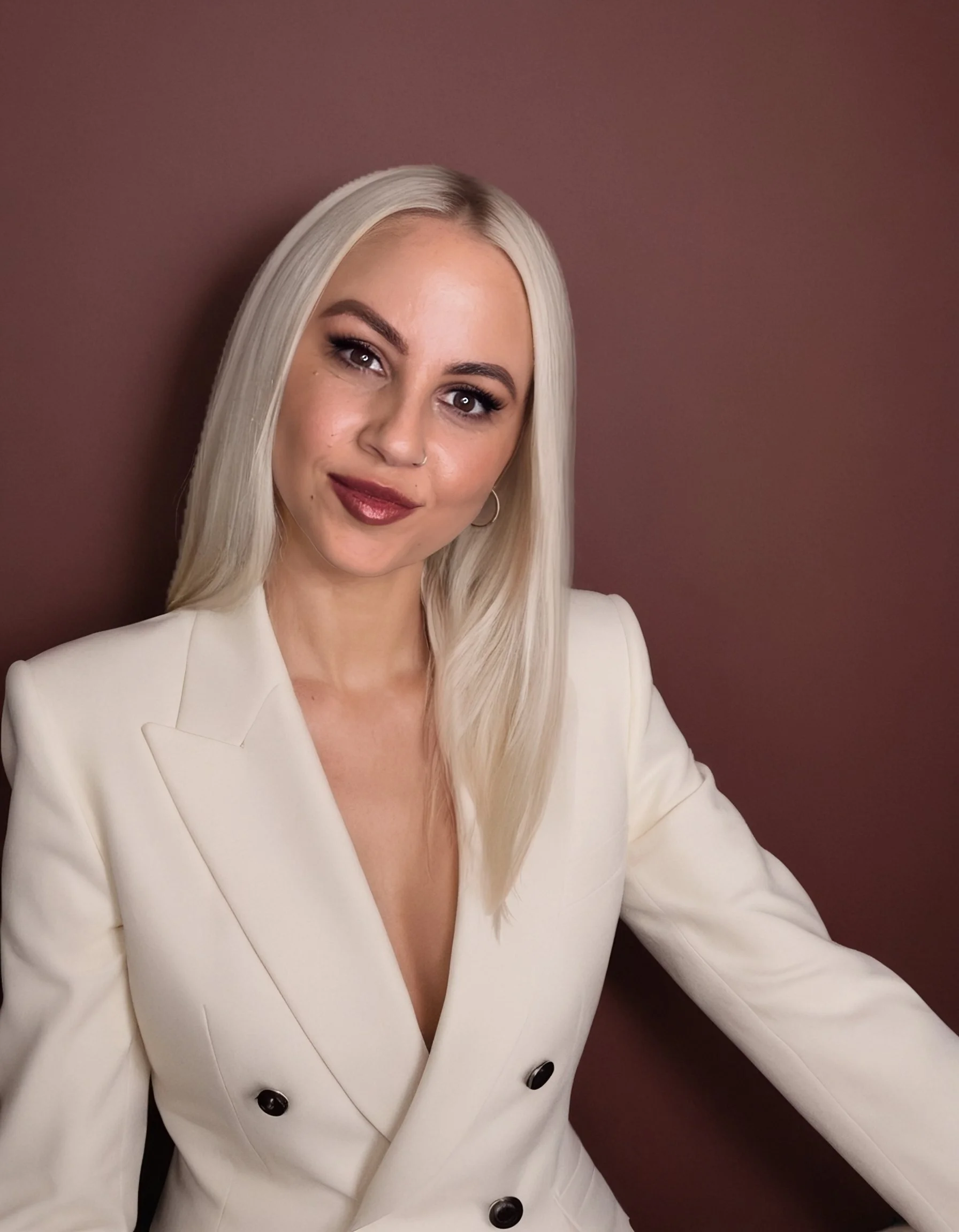 Melissa Turner, a woman with long platinum blonde hair, wearing a white blazer with black buttons, standing in front of a brown background.