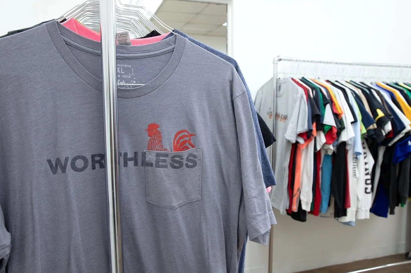 Gray vintage pocket T-shirt with a red rooster graphic and overprinted WORTHLESS lettering, hanging on a rack at WORTHLESS MARKET #1.