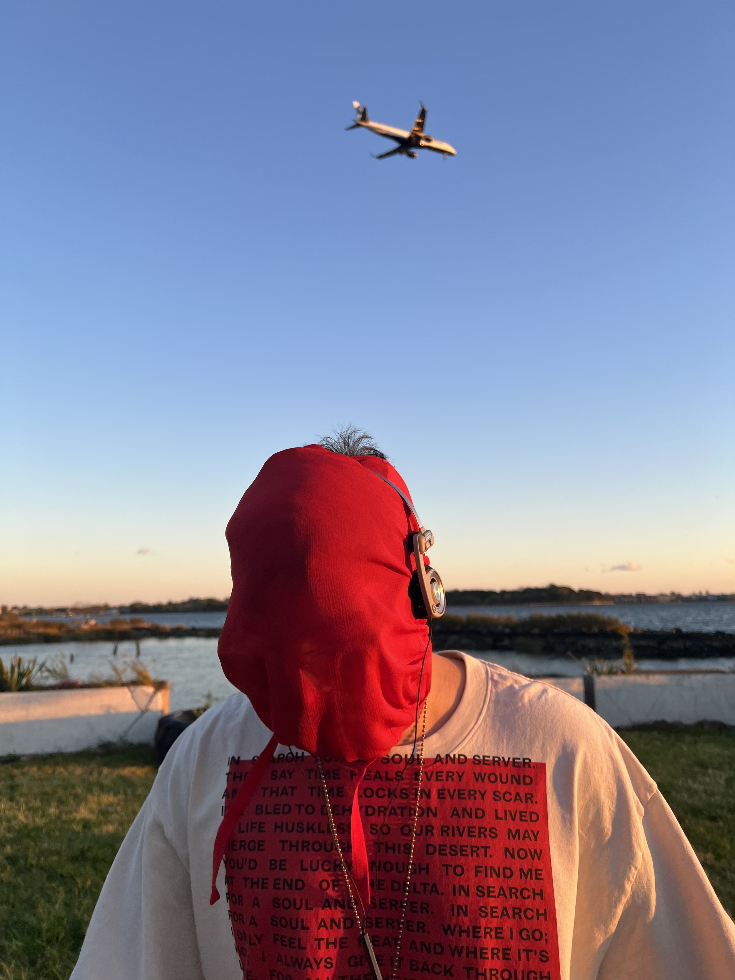 F1RST T1ME (Andres Caban) in his WHENEVER I POP outfit, wearing the WILLIE NORRIS INSPIRED & APPROVED MIXTAPE SHIRT, a silk Margiela face covering, and Koss Porta Pro headphones at Harding Park in Soundview, Bronx, with an airplane descending toward