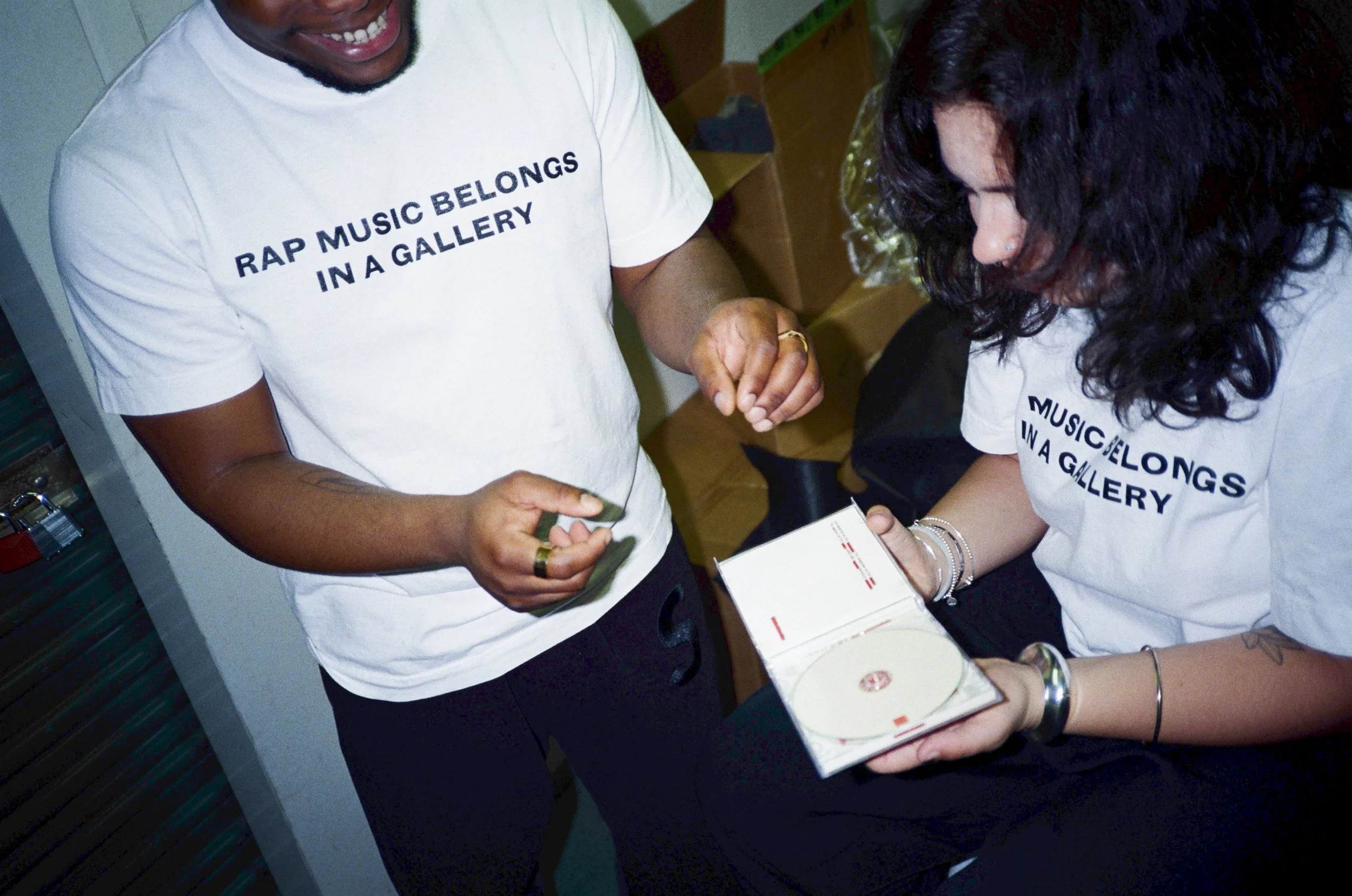 Two people wearing F1RST T1ME's white RAP MUSIC BELONGS IN A GALLERY graphic T-shirts while examining a physical CD, showing the shirt's fit and printed design.