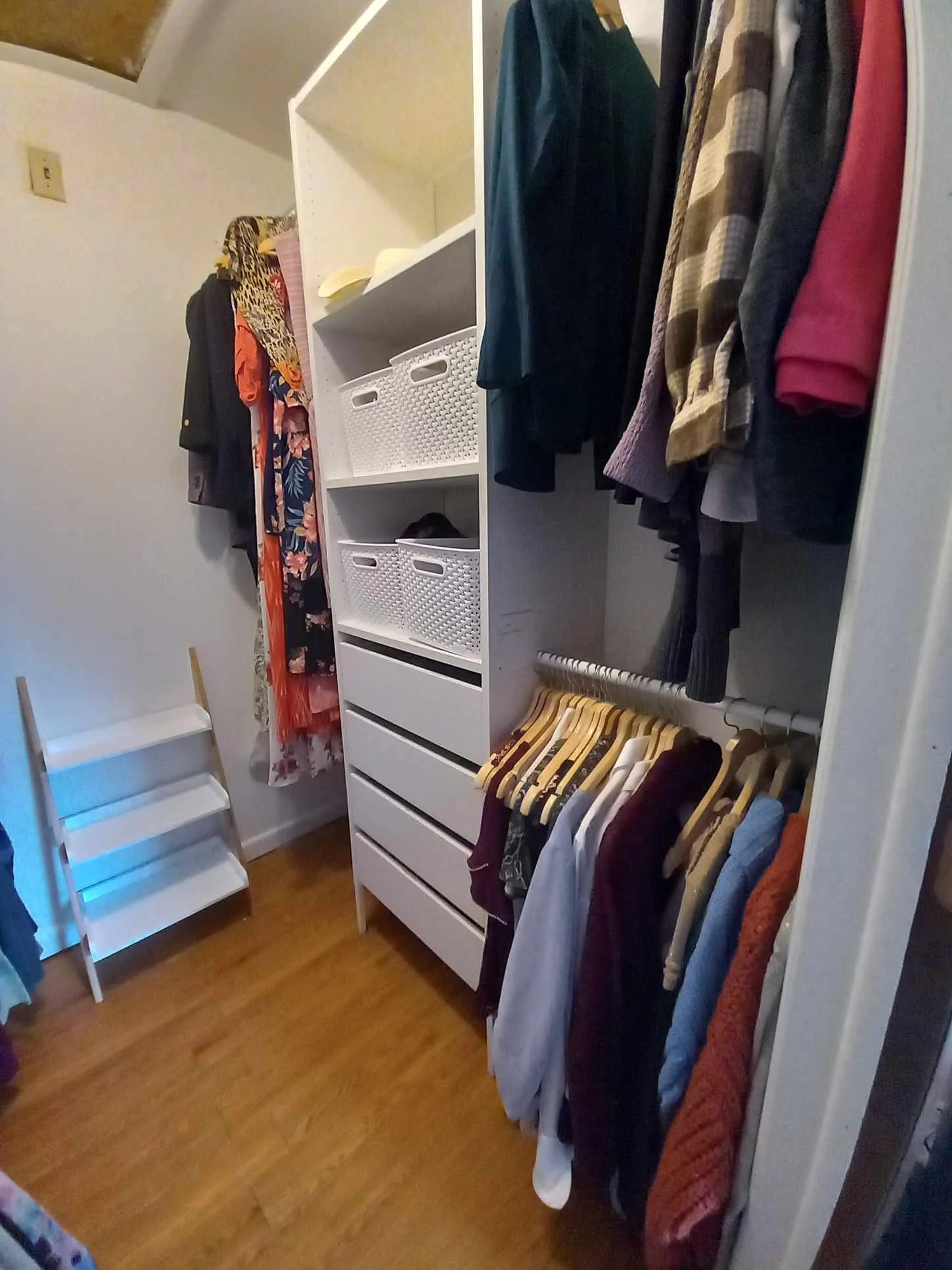 Closet with hanging clothes, white storage unit with baskets and drawers, small white stepped stool, and a wooden floor.