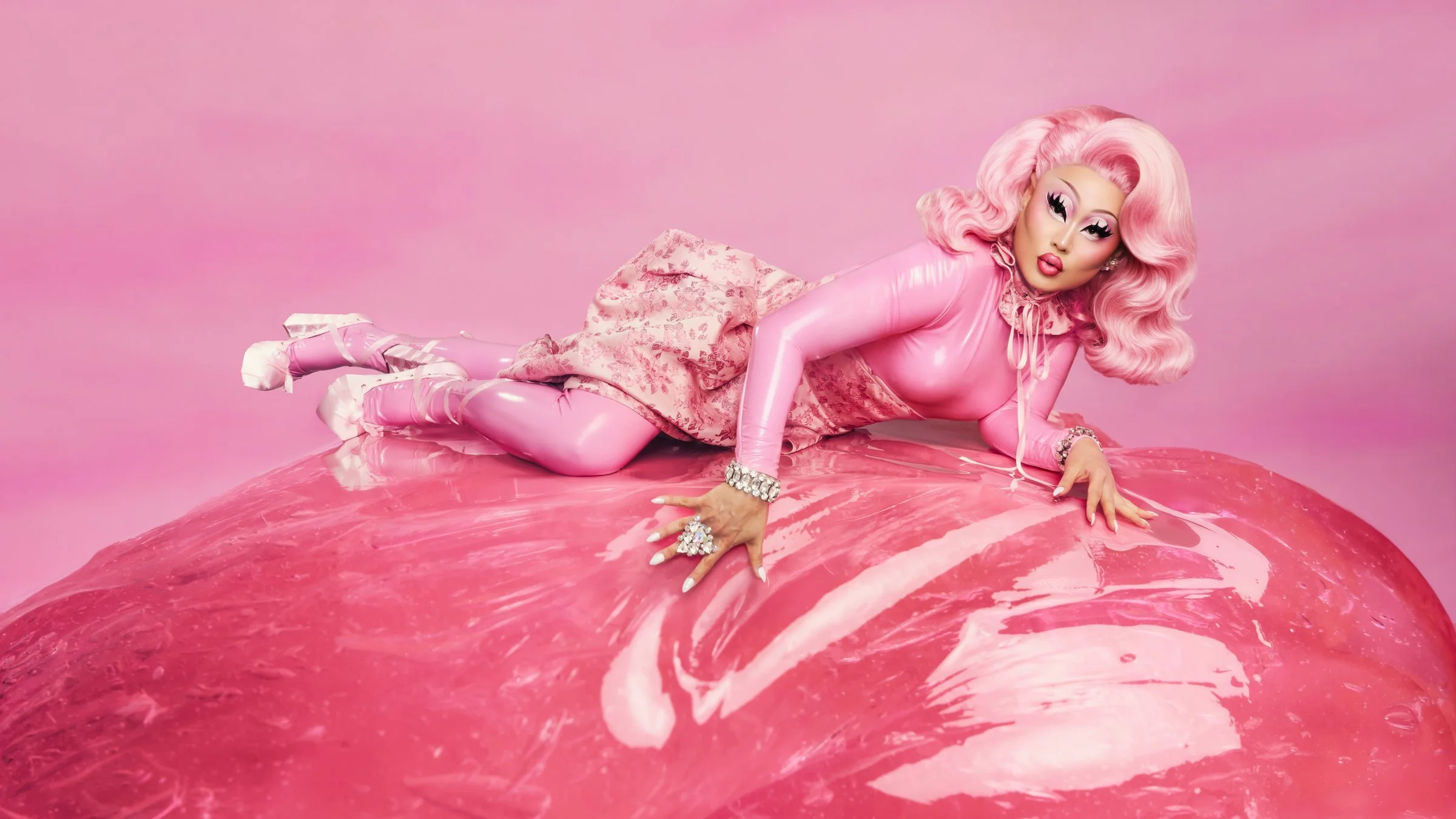 Kim Chi in a pink wig and latex outfit reclining across a glossy pink sculptural surface for KimChi Chic Beauty.