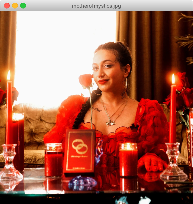 Kaitlyn Graña in a red ruffled dress holding a red rose, seated at a candlelit altar with red candles and a tarot deck