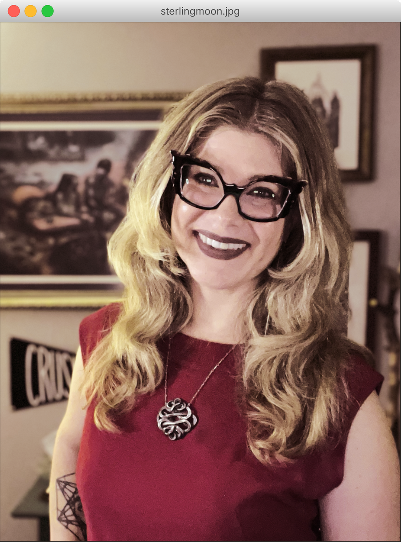 Sterling Moon smiling in cat-eye glasses and a red top in front of framed art