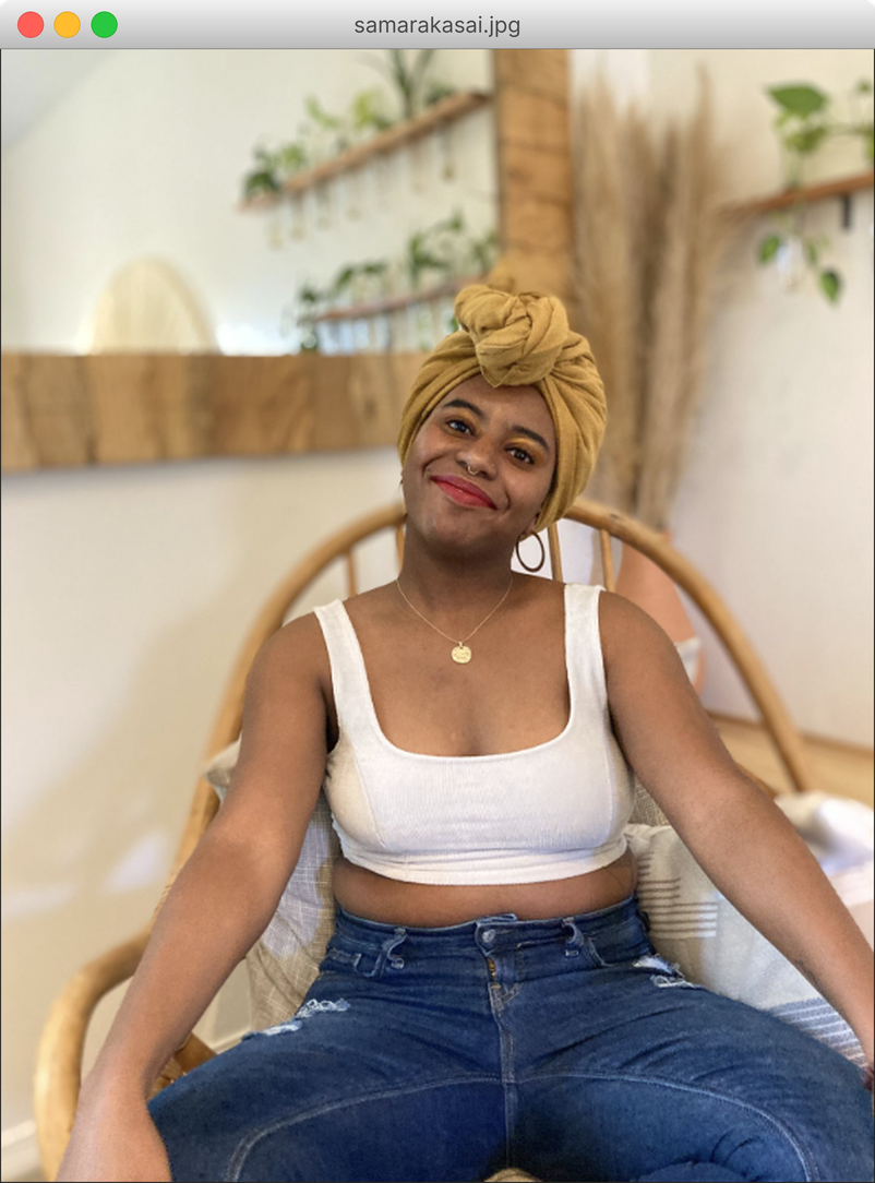 Samara Kasai smiling in a mustard head wrap, seated in a rattan chair