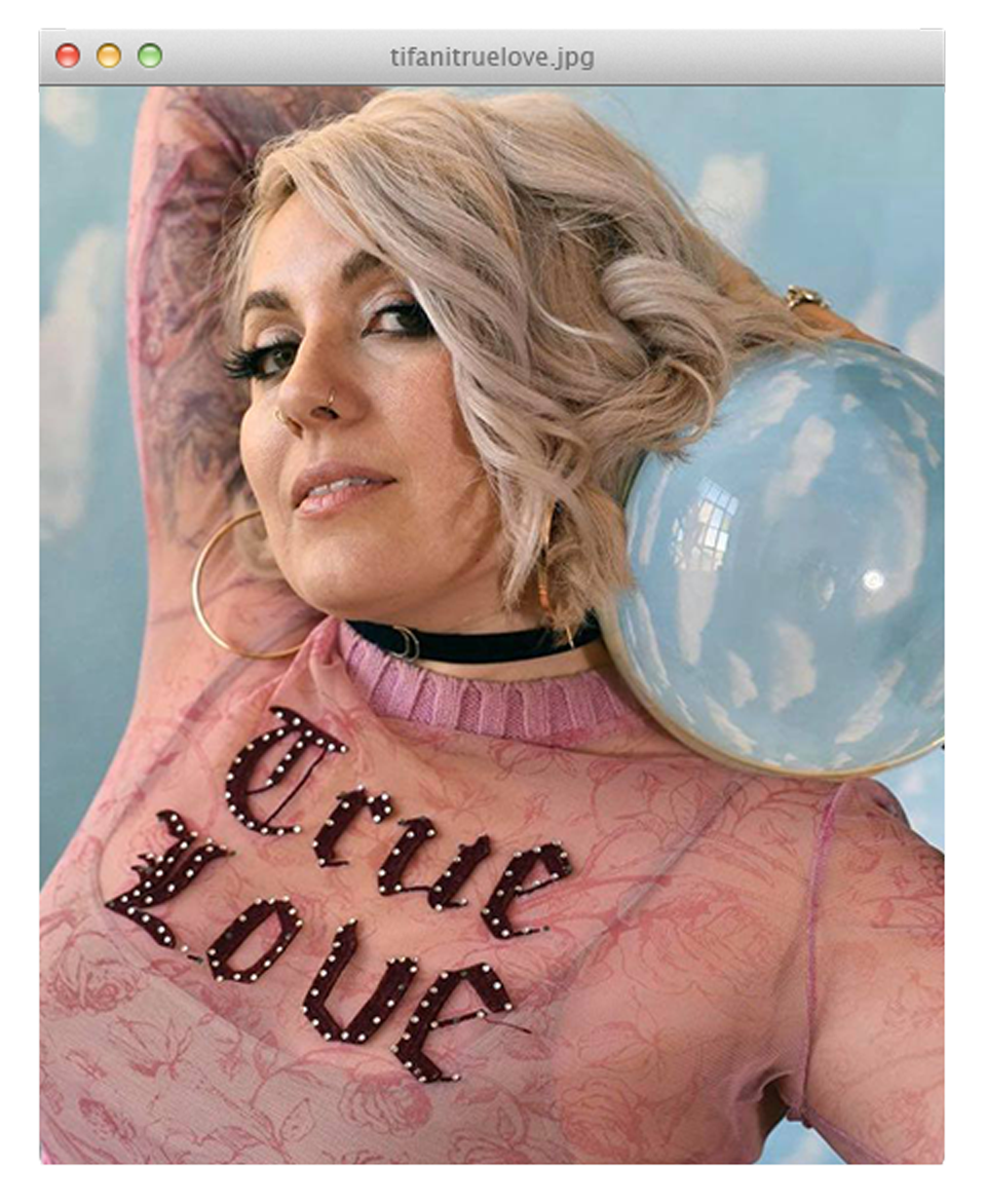 Tifani True Love in a pink sheer top reading True Love, posing with a large glass orb against a cloud backdrop