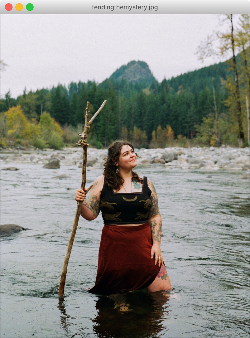 Espen Artio standing in a river holding a wooden staff, with forest and mountains behind