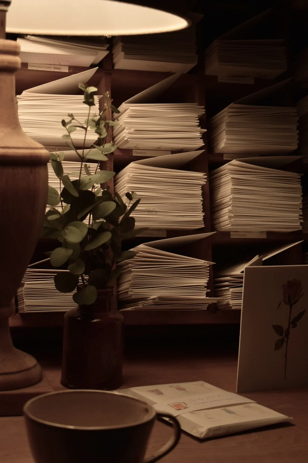 A desk with a cup, a brown vase with green leaves, open card and book, set against a background of wooden shelves filled with neatly stacked papers or notebooks.