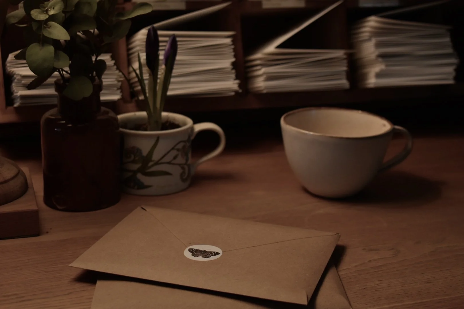 A wooden desk with a plant in a brown vase, a cup with a floral pattern holding budding flowers, an empty ceramic cup, and a closed envelope with a butterfly sticker, against a backdrop of organized shelves with papers.