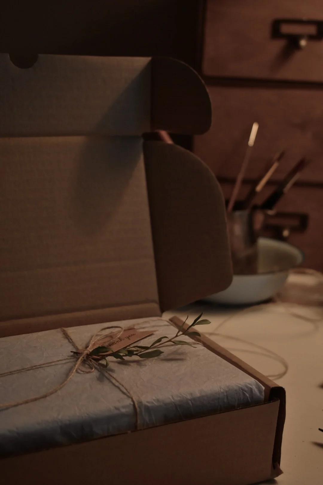 A cardboard box with wrapping paper and a sprig of greenery on top, placed on a table with a dimly lit background including a wooden cabinet and some utensils.