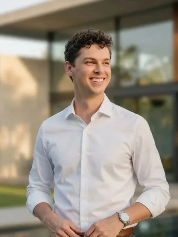 A young man with curly dark hair and a white dress shirt smiling outdoors with a modern building in the background.
