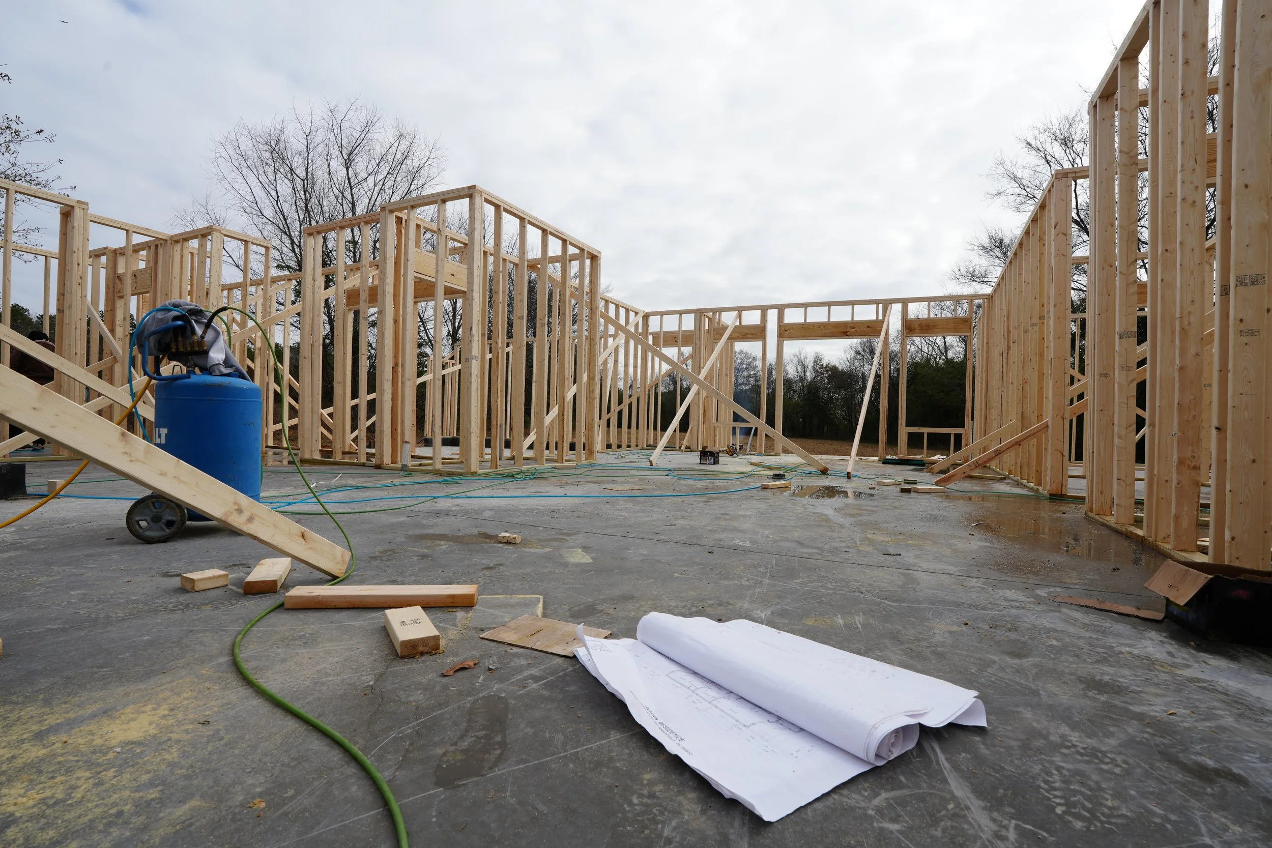 Construction site with incomplete wooden framing for a building. Tools and construction plans are on the ground, and a blue air compressor is visible. Trees and a cloudy sky are in the background.