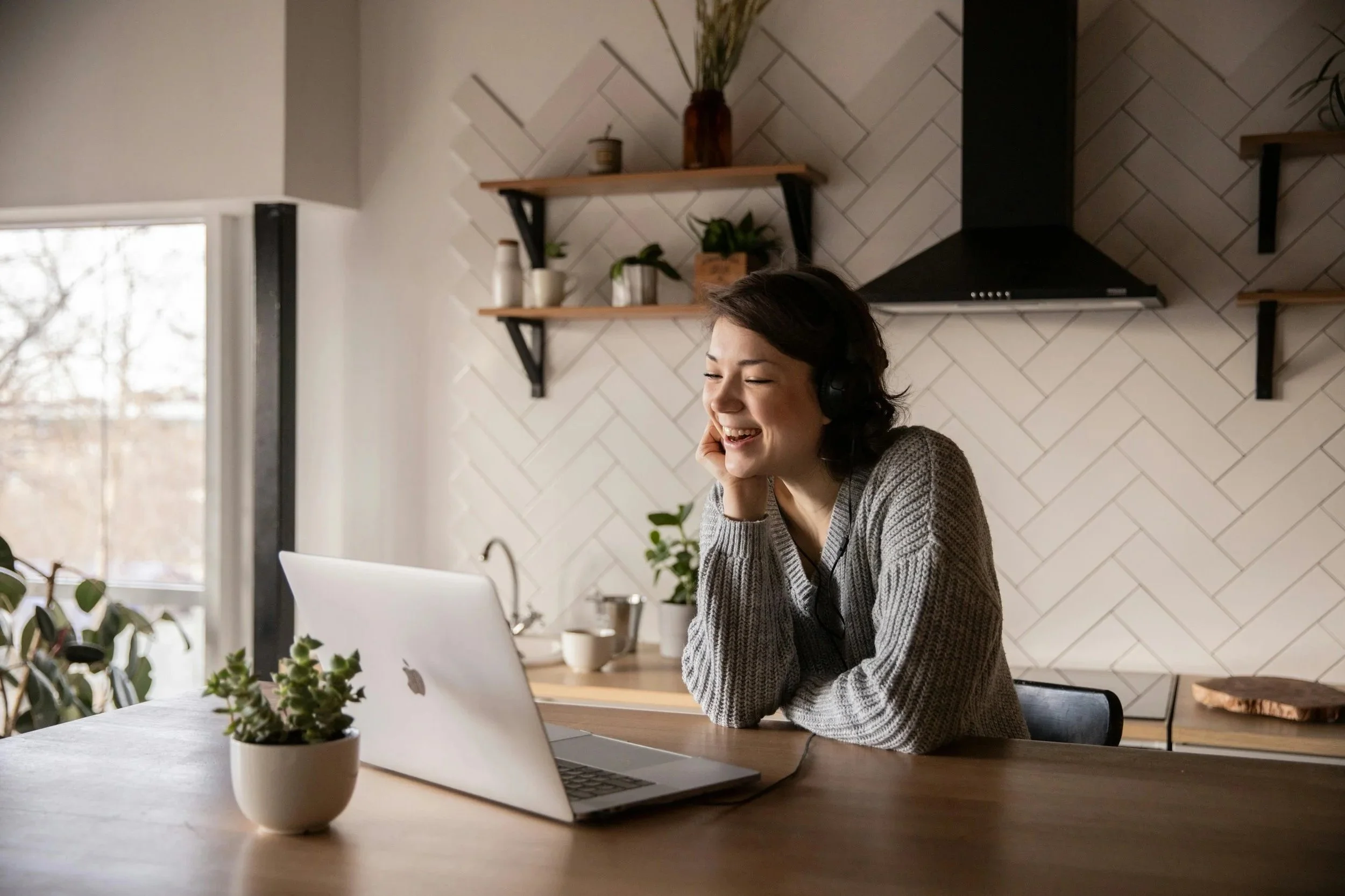 A woman with dark hair, wearing a gray sweater, smiling and wearing headphones, sitting at a kitchen table with a laptop, a potted plant, and other kitchen items in the background.