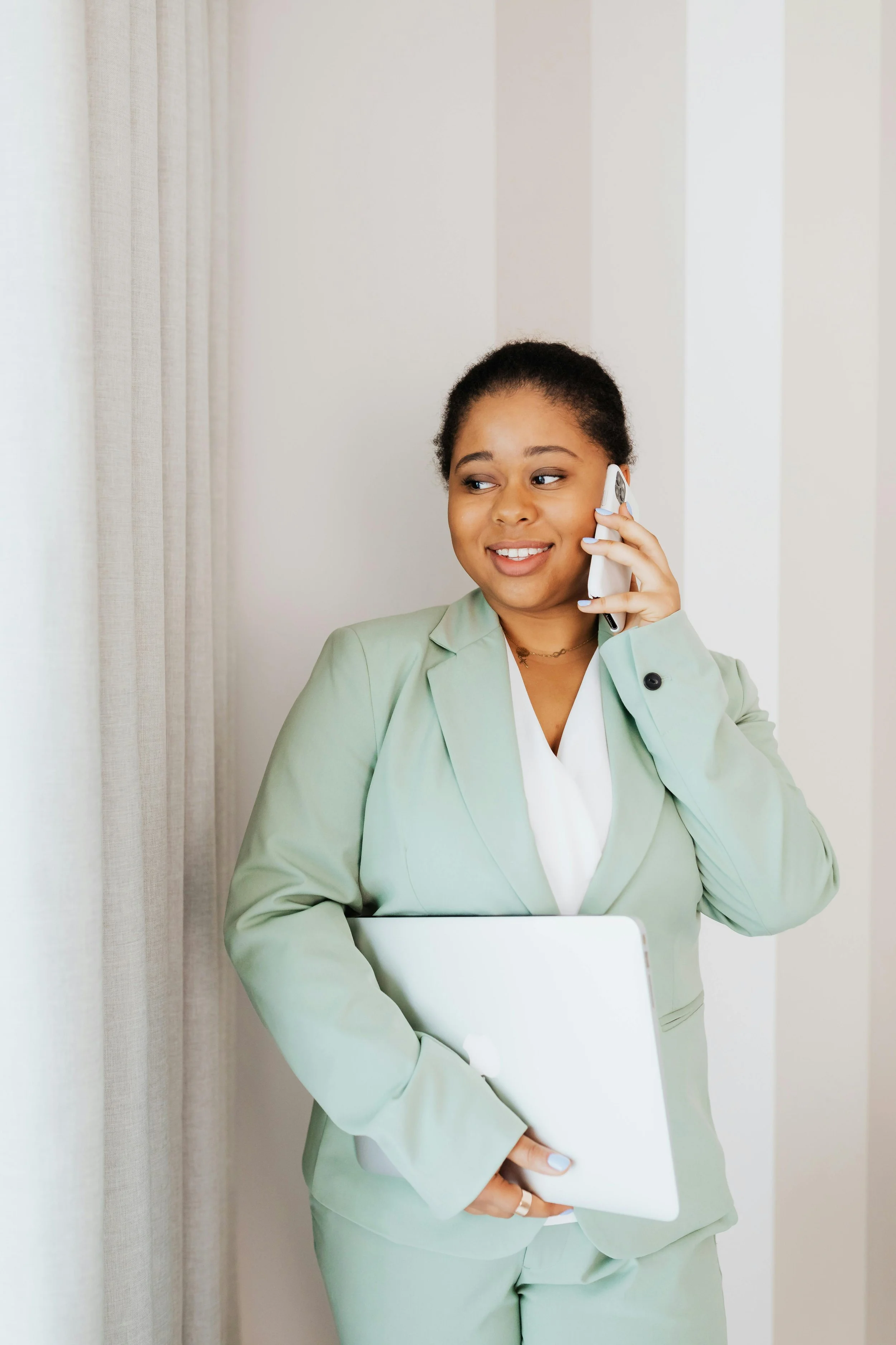 A woman in a light green suit holding a closed laptop and talking on a cordless phone, standing near a curtain in a bright room.