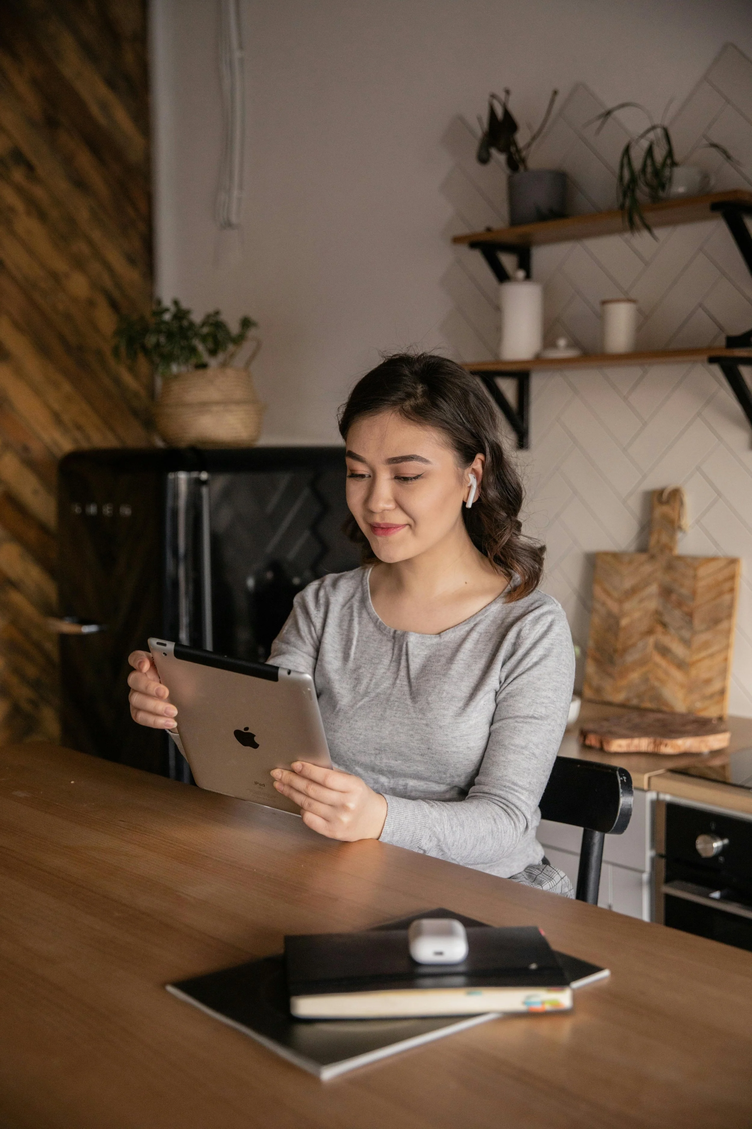 A young woman with dark hair, wearing a gray long-sleeve shirt and white wireless earbuds, is sitting at a wooden table, holding an iPad, smiling slightly. On the table in front of her, there are a notebook, a closed black folder, and a pair of white wireless earbuds case. The background shows a white wall with wooden shelves, decorative plants, and kitchen items.