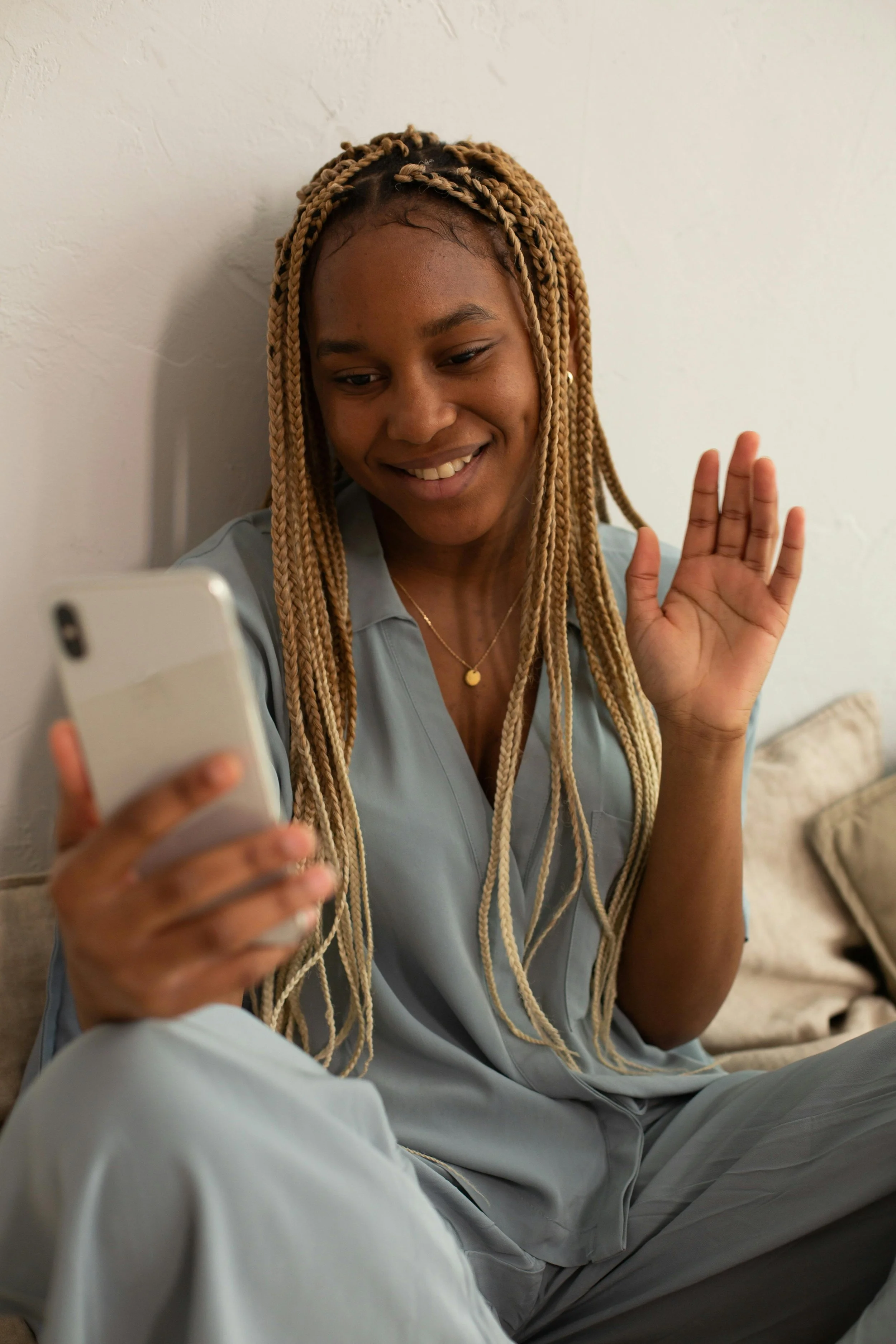 A woman with long blonde braids smiling and waving while looking at her phone, sitting on a beige couch against a white wall.