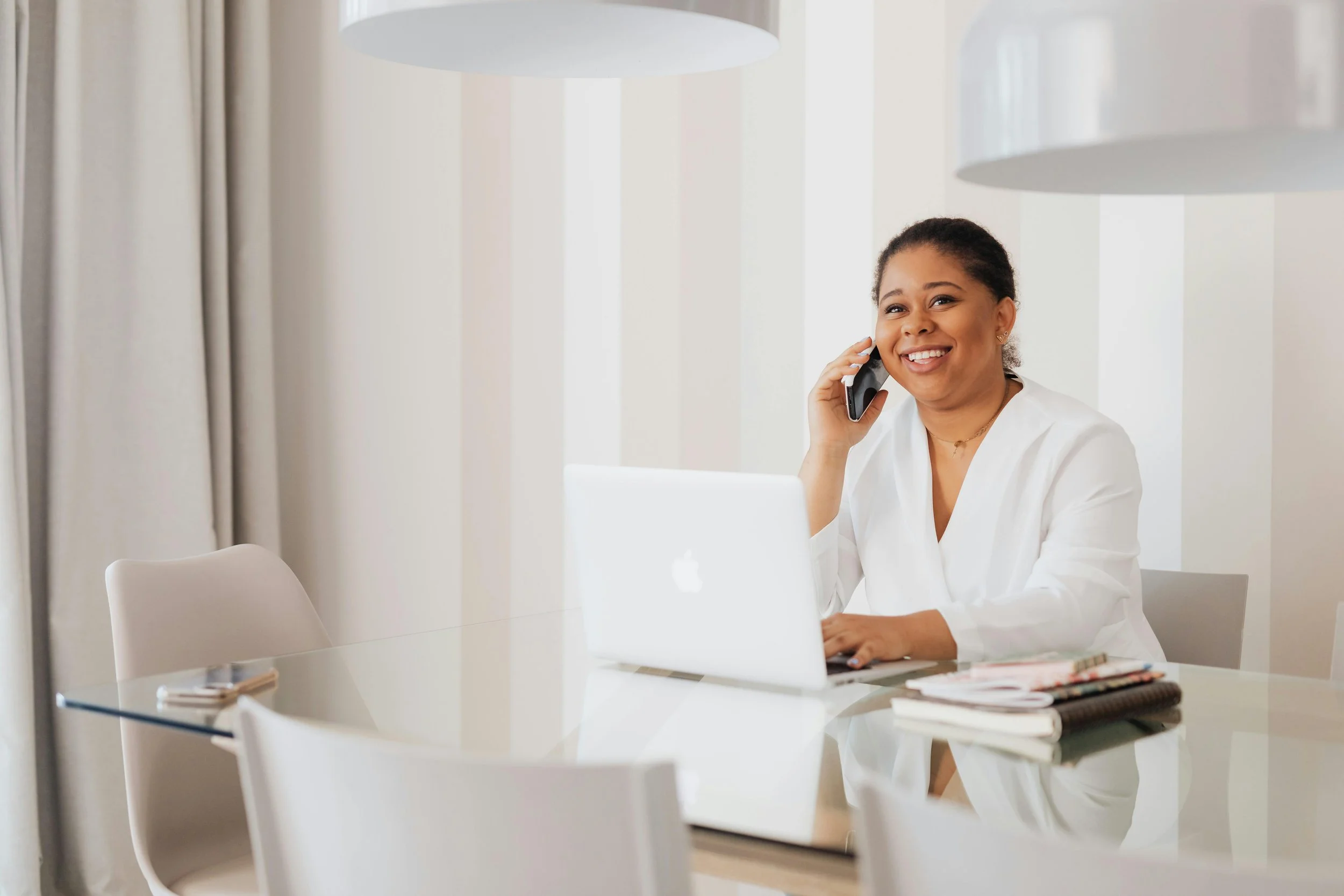 Woman smiling and talking on phone at desk with laptop and notebooks in a bright room.