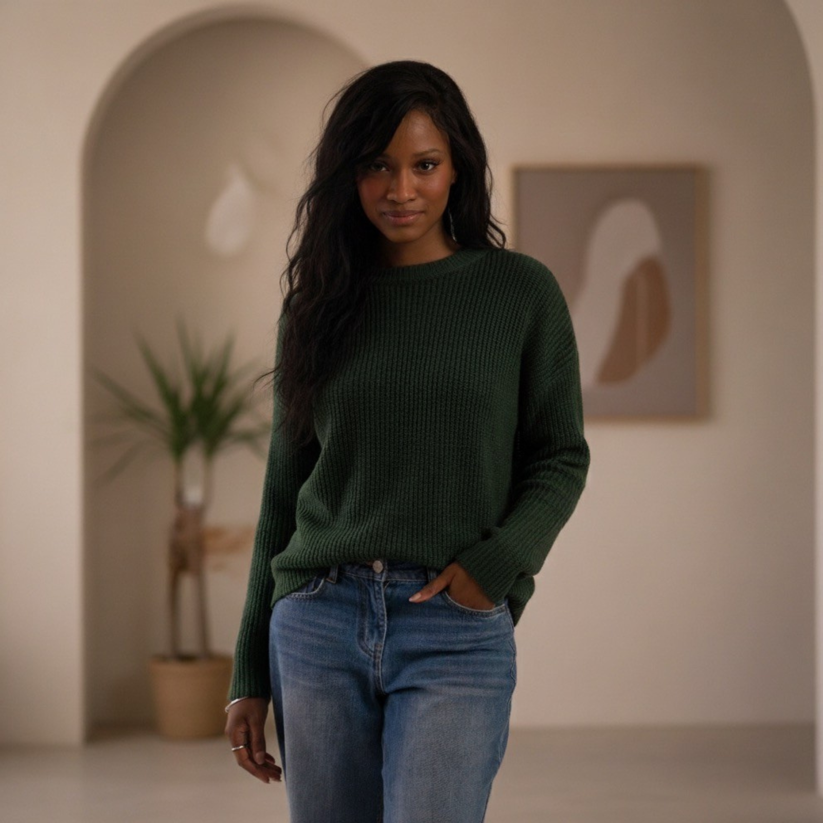 O'Shea René with dark wavy hair wearing a green sweater and blue jeans standing indoors with a plant and abstract artwork in the background.