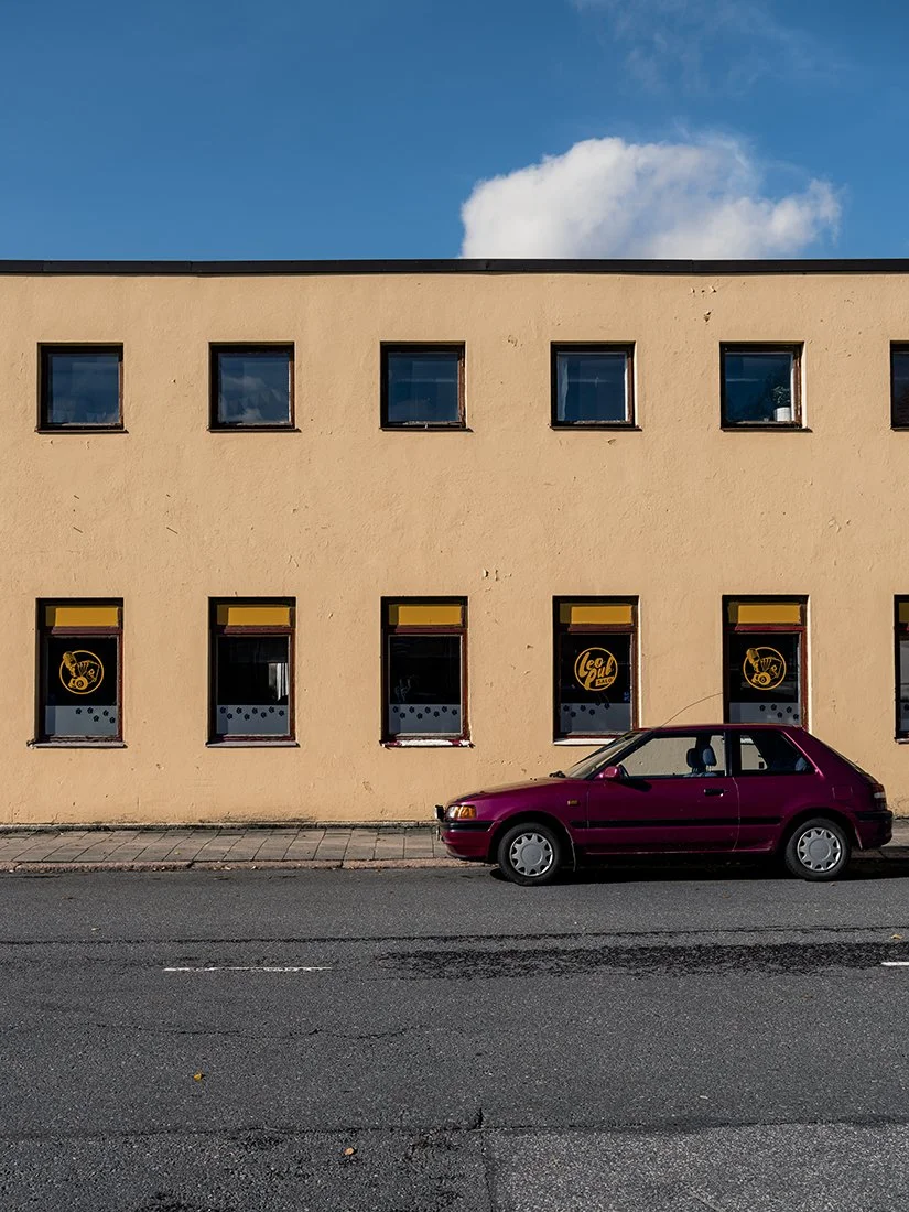 A red car parked on a street in front of a beige building with six windows. The building has a flat roof, and the sky is blue with some clouds.