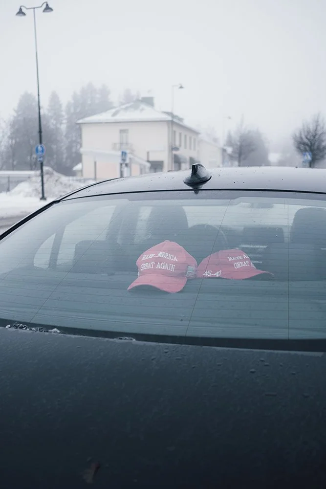 Two red 'Make America Great Again' hats on the rear window of a car, with a snowy outdoor scene and a building in the background.
