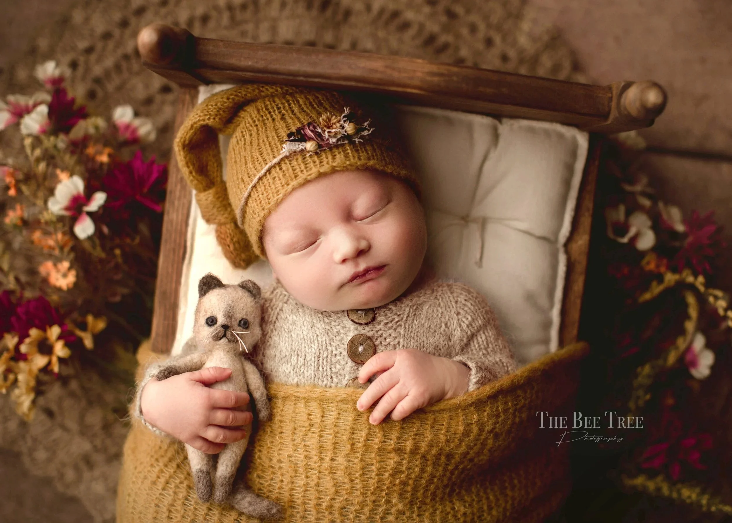 A sleeping baby wearing a brown knitted hat with a decorative flower, lying in a small wooden bed with a white pillow wrapped in a brown knitted blanket. The baby is holding a small felted animal, possibly a mouse or a bear, and is dressed in a beige