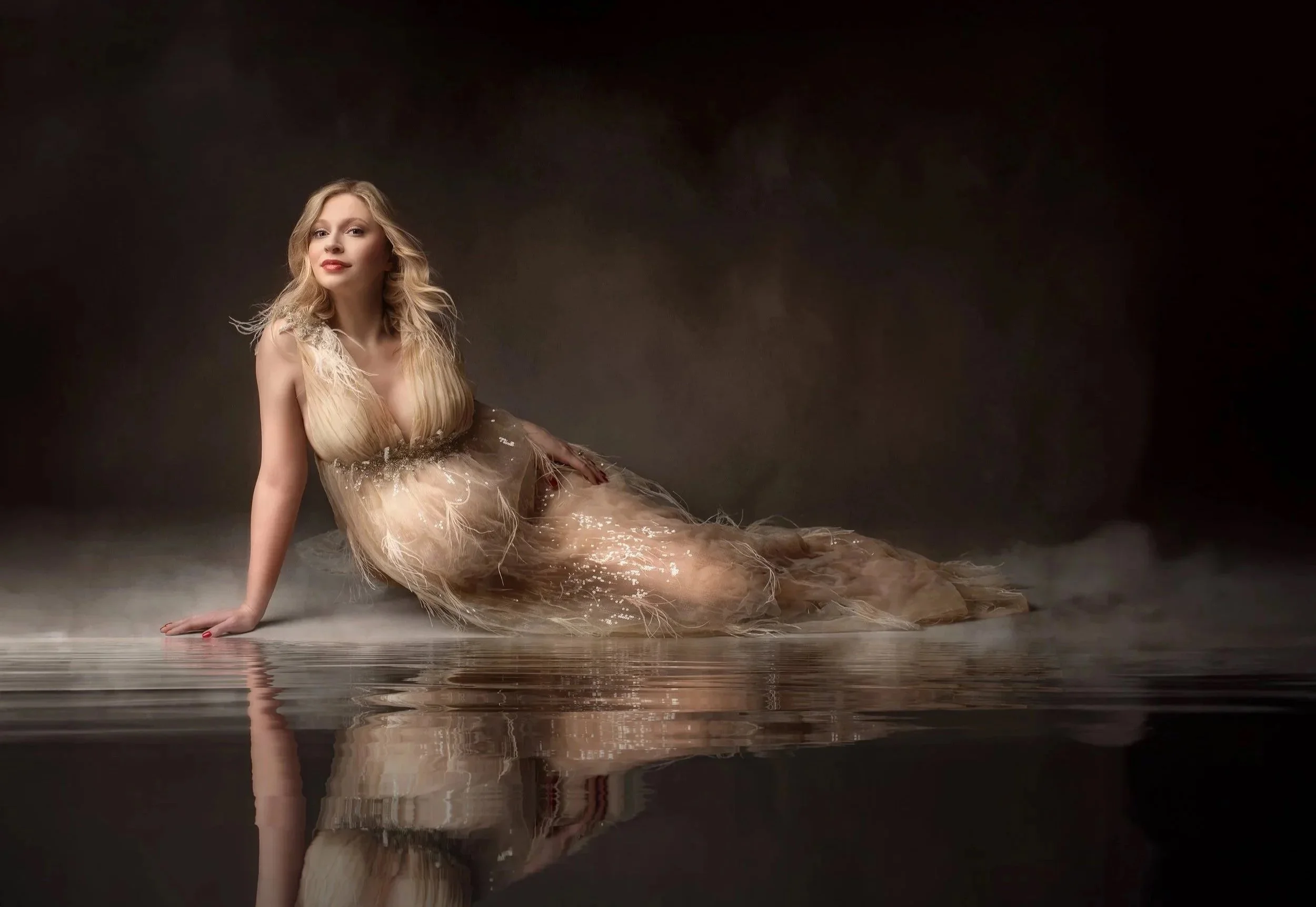 A woman with long blonde hair sitting on a reflective surface in a flowing, tan-colored dress with feather details, against a dark background.