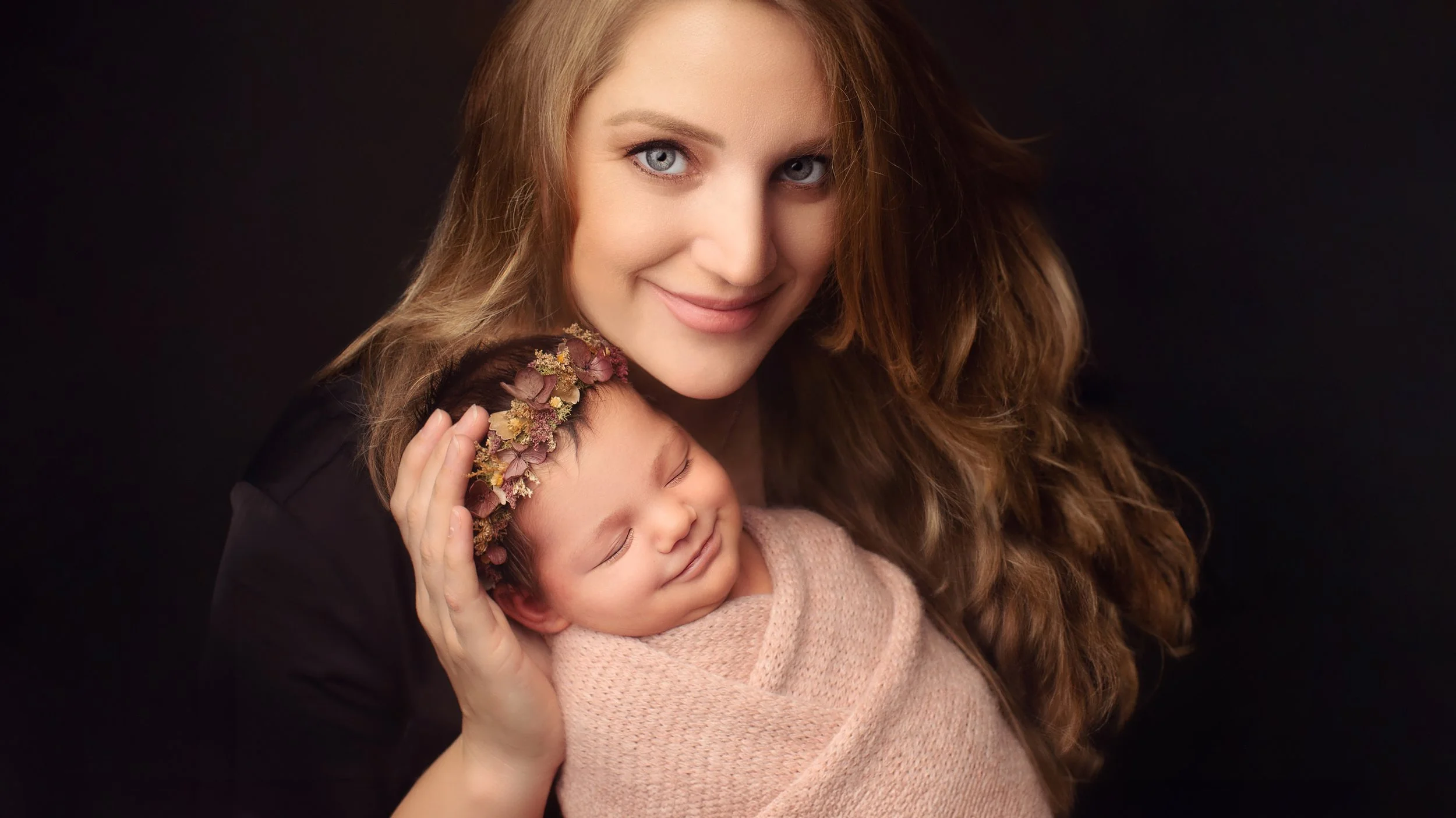 A woman with long wavy hair holding a sleeping baby wrapped in a pink blanket, wearing a floral headband, with a dark background.