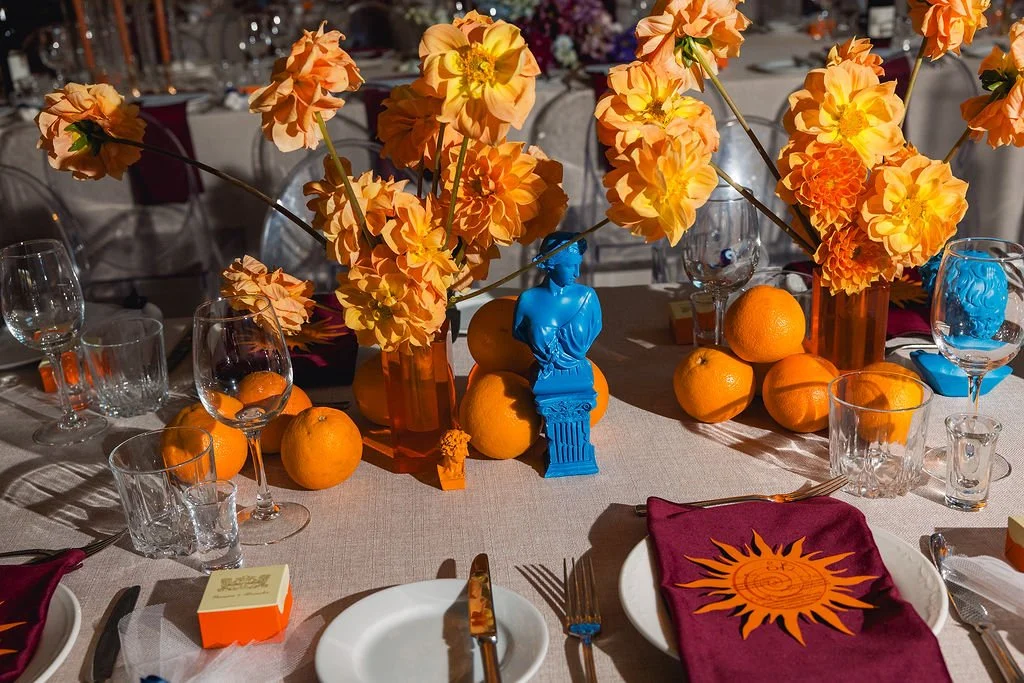 ornage dahlias placed in en masse in orange retro style glass vases. featured with electriv blue busts. the vases are linked with oranges whole. the table is dressed with natural coloured linen, deep burgandy napkins and the menus are orange suns