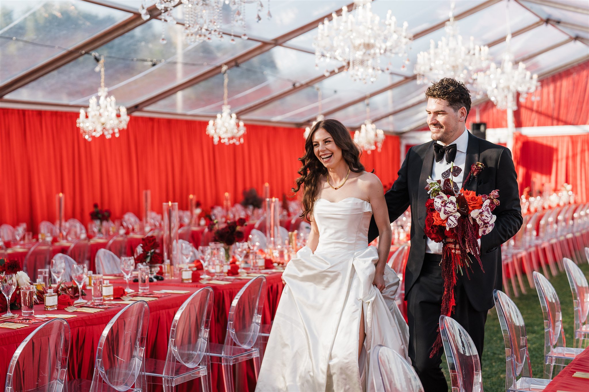 Renee and Austin smile as they walk through their reception prior to guests. The Marquee has a clear top and is dressed with red velvet draped walls, clear glass chandeliers, matching red velvet table cloths and clear ghost chairs
